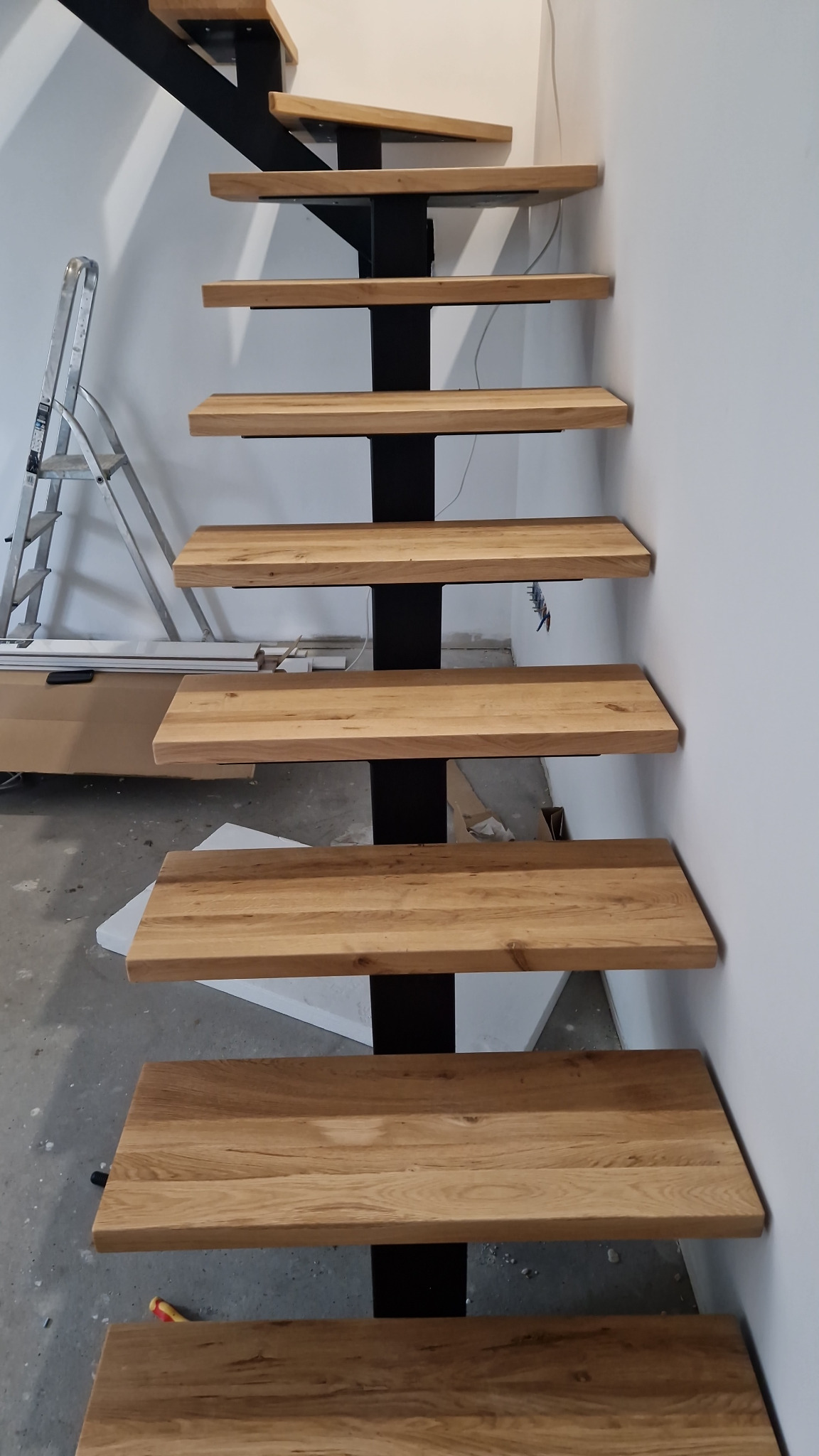 Drewniane stopnie schodów na czarnej, metalowej konstrukcji, widoczne niedokończone prace wykończeniowe i drabina malarska w tle.