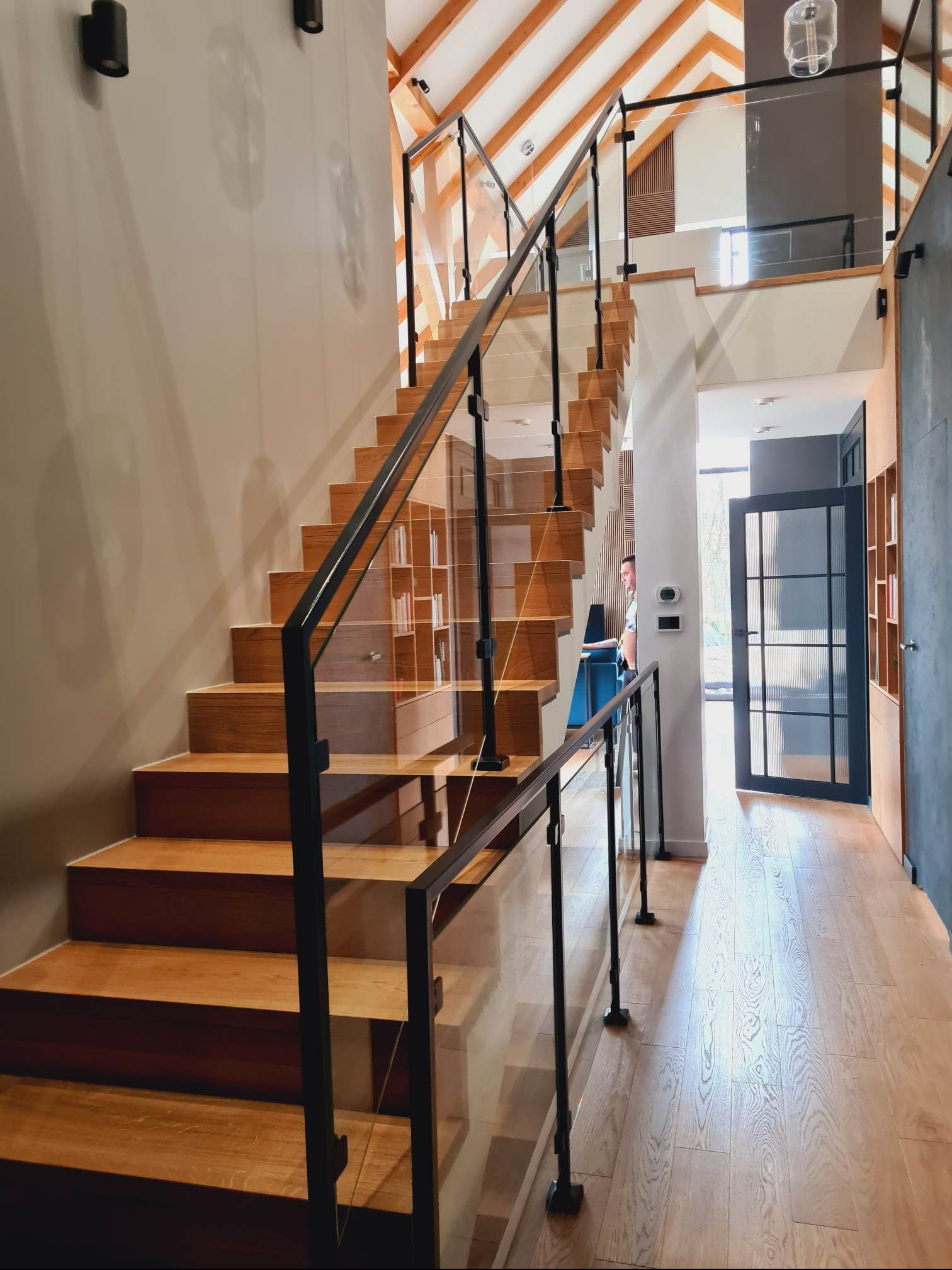 Nowoczesne, drewniane schody z czarną balustradą i szklanymi panelami w jasnym wnętrzu domu, widok z dołu na górę, drewniana podłoga, drzwi z przeszkleniami w tle.