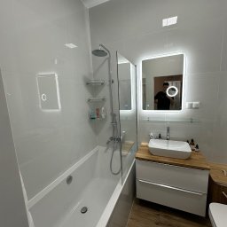 Usługi Remontowo-Budowlane Wojciech Kudłaciak EKO-BUDOWA - Nowoczesna łazienka z wanną, przeszklonym parawanem nawannowym, umywalką nablatową na drewnianym blacie i podświetlanym lustrem; odbicie fotografa w lustrze.