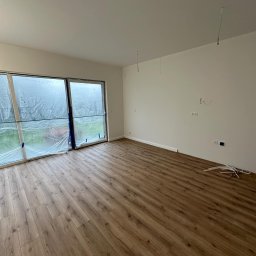 Usługi Remontowo-Budowlane Wojciech Kudłaciak EKO-BUDOWA - Pokój po remoncie z panelami podłogowymi w odcieniach brązu, okna zabezpieczone folią malarską, wystające kable ze ściany i sufitu, oraz listwy przypodłogowe czekające na montaż.