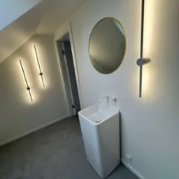 Nowoczesna łazienka na poddaszu z wolnostojącą, kwadratową umywalką, okrągłym lustrem w złotej ramie i minimalistycznymi, pionowymi lampami LED.