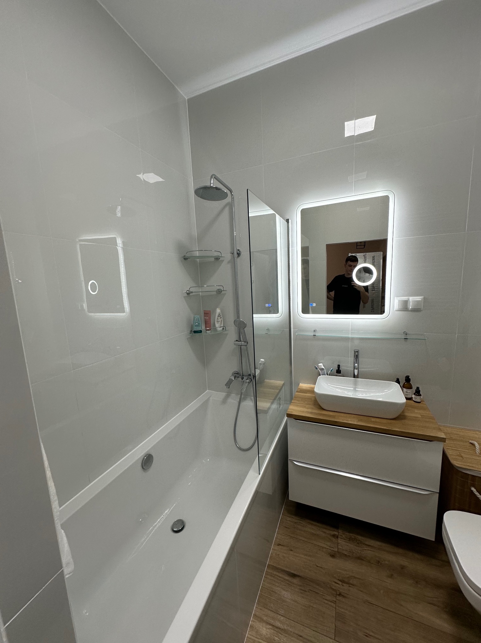 Nowoczesna łazienka z wanną, przeszklonym parawanem nawannowym, umywalką nablatową na drewnianym blacie i podświetlanym lustrem; odbicie fotografa w lustrze.