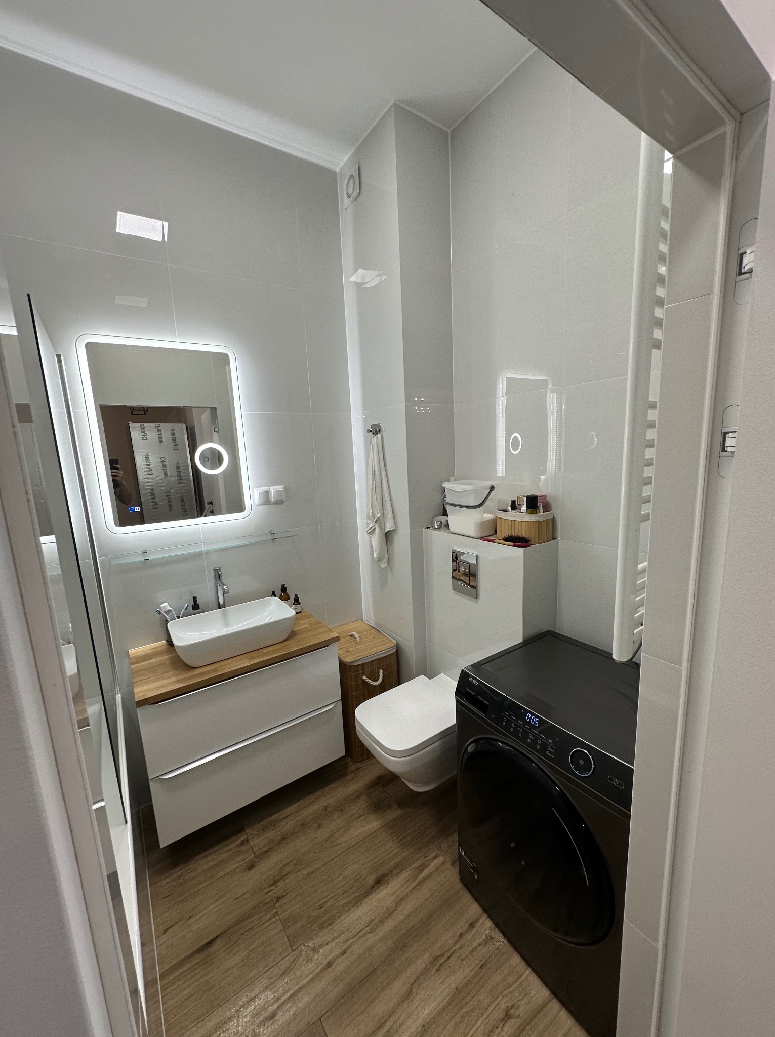 Nowoczesna łazienka z białymi płytkami, drewnopodobną podłogą, umywalką nablatową na drewnianym blacie, podświetlanym lustrem, toaletą i czarną pralką Haier.
