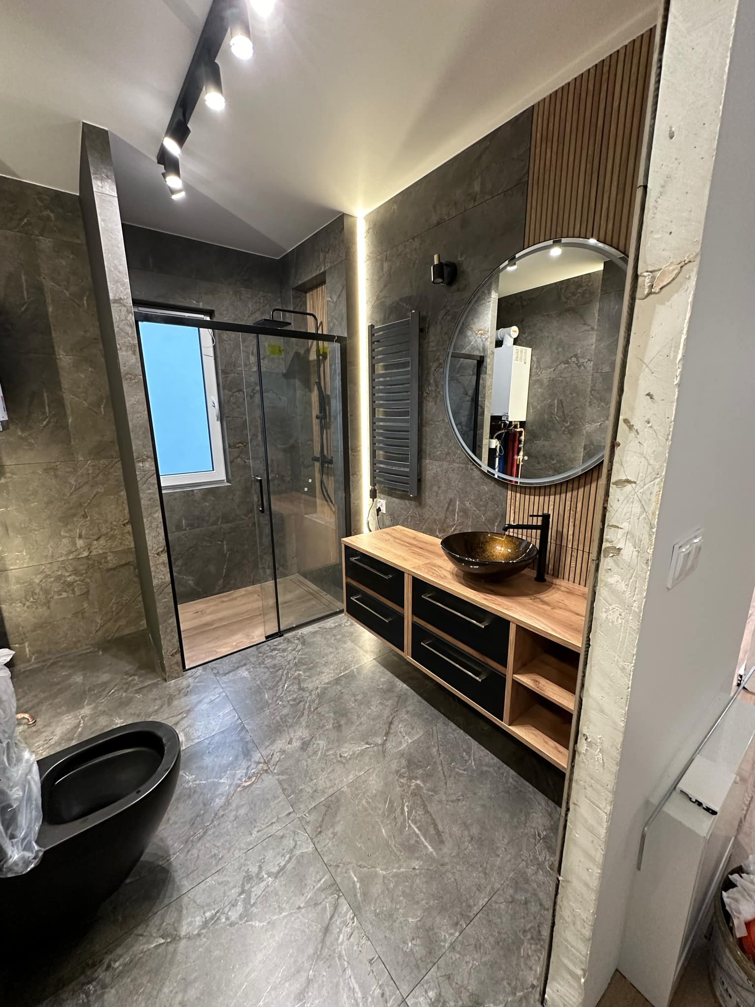 Nowoczesna łazienka z kabiną prysznicową typu walk-in, umywalką w kolorze bursztynu na szafce z drewnianym blatem i czarnymi frontami, okrągłym lustrem i grzejnikiem drabinkowym na tle szarych...