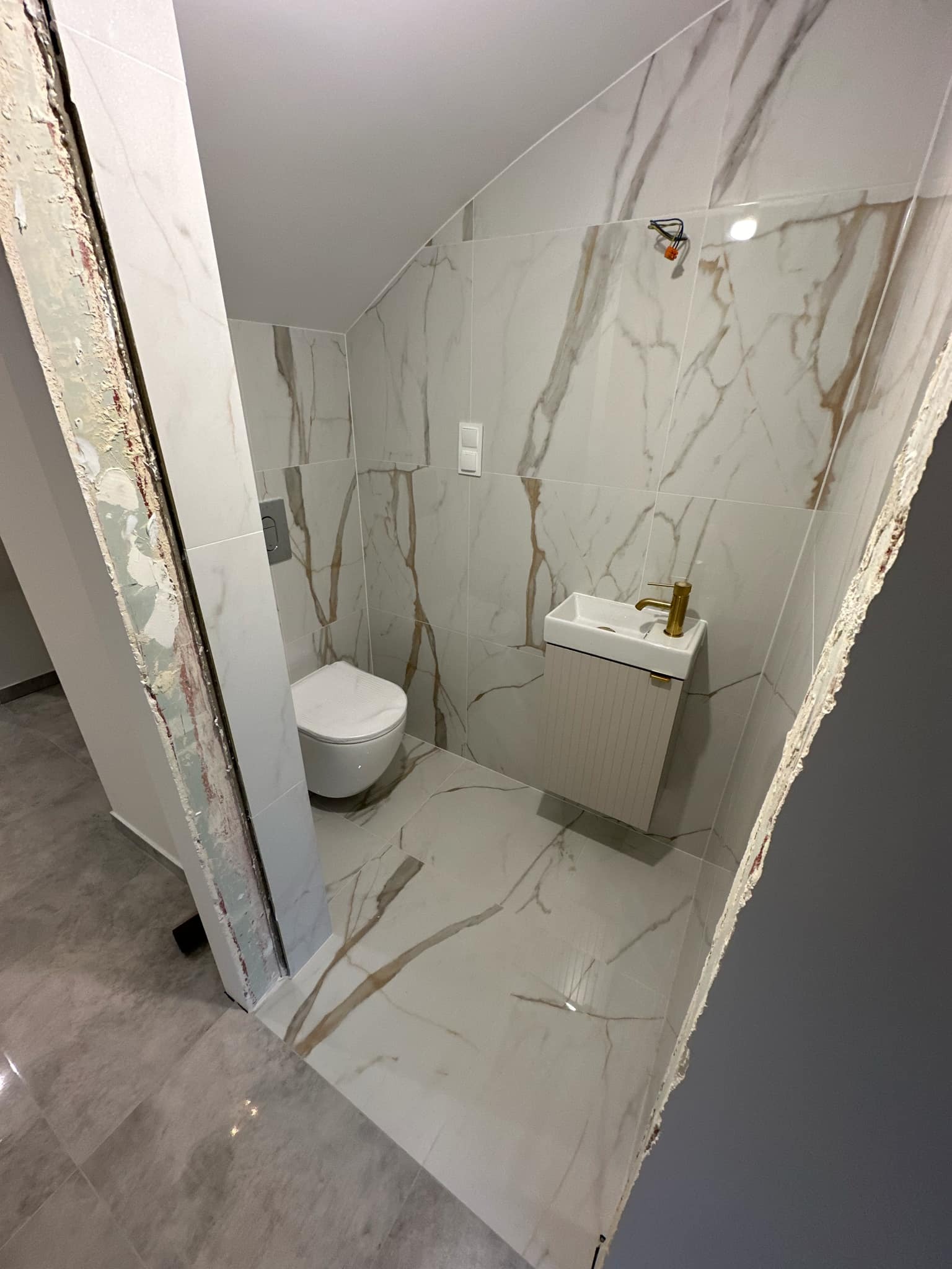 Mała łazienka z białymi płytkami imitującymi marmur, wisząca toaleta i szafka z umywalką z baterią w kolorze złotym, widoczne niedokończone obramowanie drzwi.
