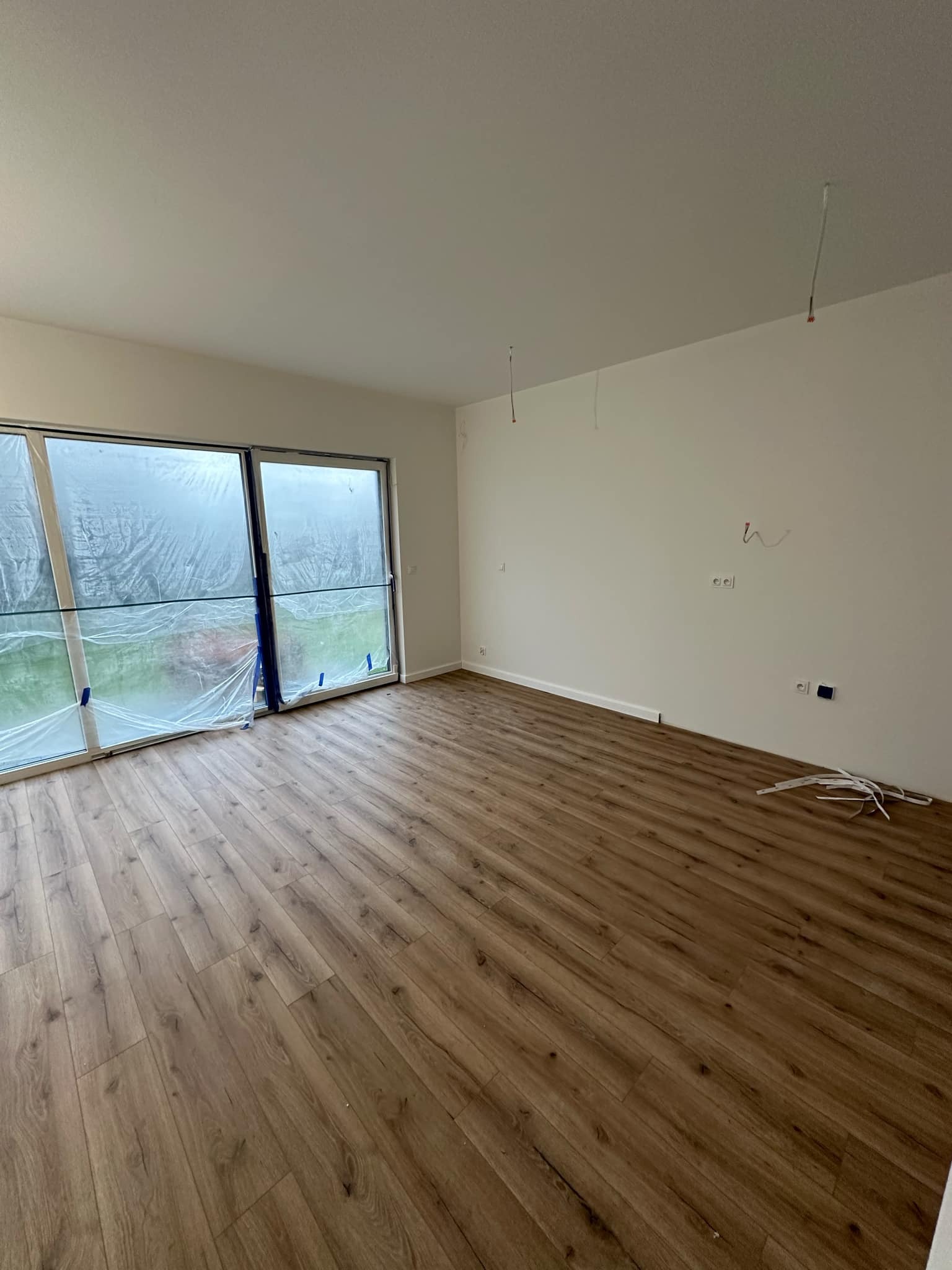 Pokój po remoncie z panelami podłogowymi w odcieniach brązu, okna zabezpieczone folią malarską, wystające kable ze ściany i sufitu, oraz listwy przypodłogowe czekające na montaż.