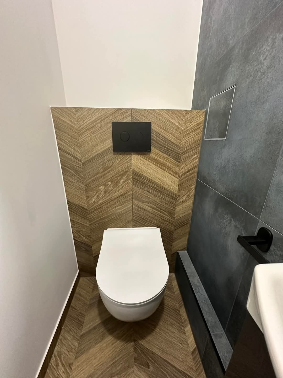 Nowoczesna toaleta podwieszana z zabudową z drewnopodobnych płytek ułożonych w jodełkę, czarny przycisk spłuczki i szara ściana z odpływem liniowym.
