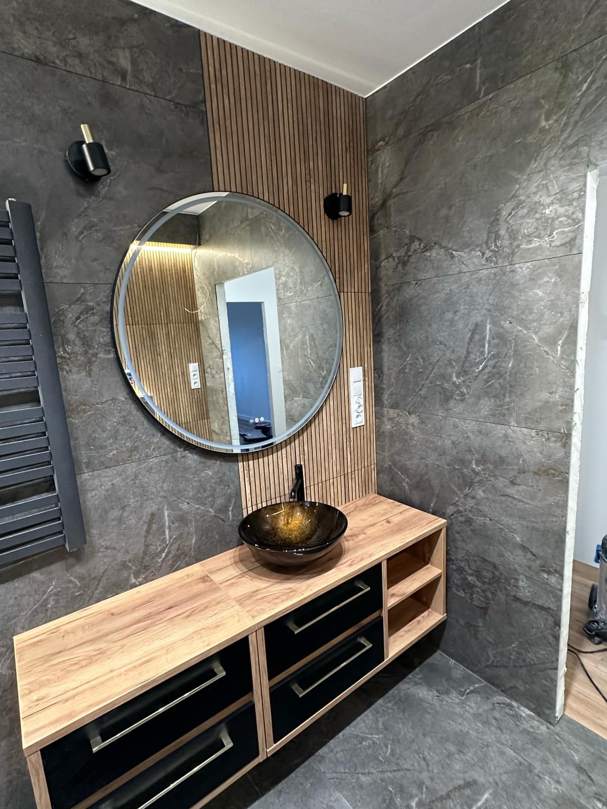 Nowoczesna łazienka w Gdańsku z umywalką w odcieniach brązu, okrągłym lustrem z podświetleniem LED, drewnianymi lamelami na ścianie i szarymi płytkami imitującymi marmur.