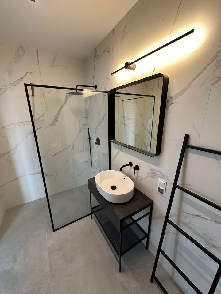 Nowoczesna łazienka z kabiną prysznicową typu walk-in z czarnymi ramami, umywalką nablatową na czarnym postumencie, lustrem w czarnej ramie i drabinką na ręczniki, wykończona marmurowymi płytkami.