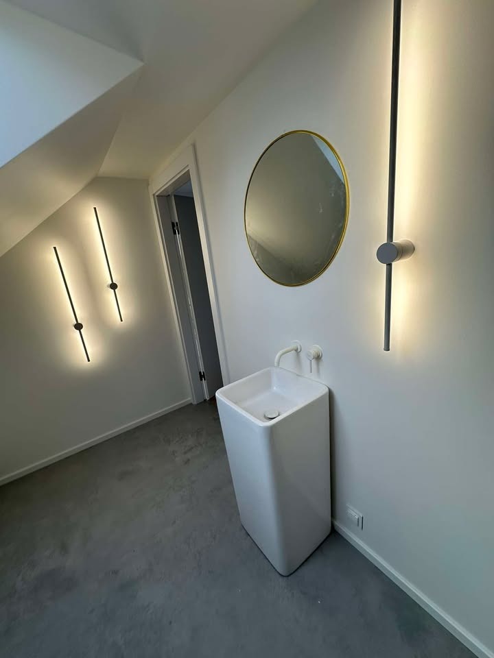 Nowoczesna łazienka na poddaszu z wolnostojącą, kwadratową umywalką, okrągłym lustrem w złotej ramie i minimalistycznymi, pionowymi lampami LED.