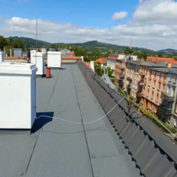 Widok z góry na płaski, szary dach pokryty papą, z białymi kominami i anteną, w tle zabudowa miejska z budynkami o pomarańczowych dachach i zielenią drzew.