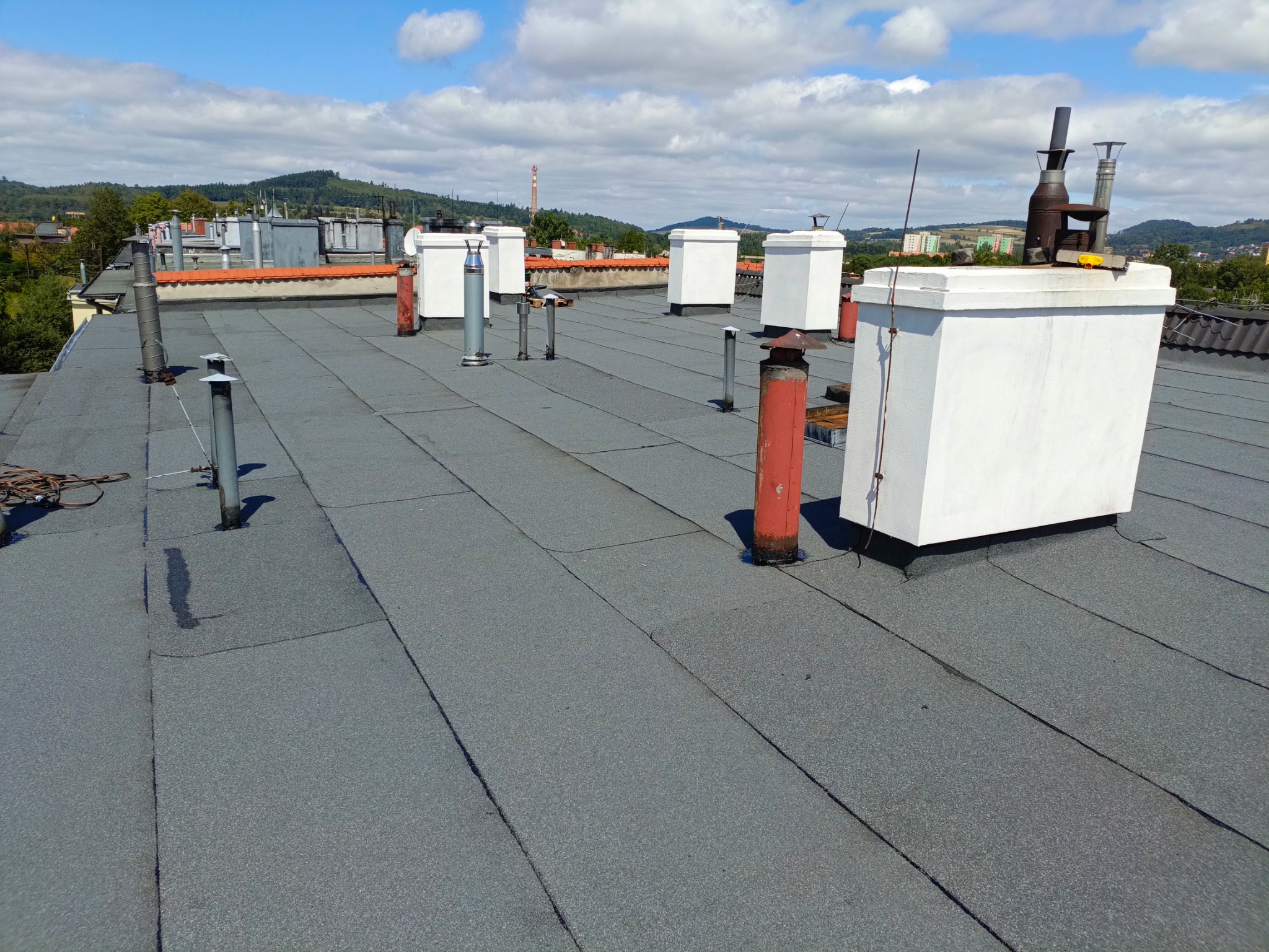 Widok na płaski dach pokryty papą termozgrzewalną z kominami i wentylacją, w tle zabudowa miejska i zalesione wzgórza pod błękitnym niebem z chmurami.