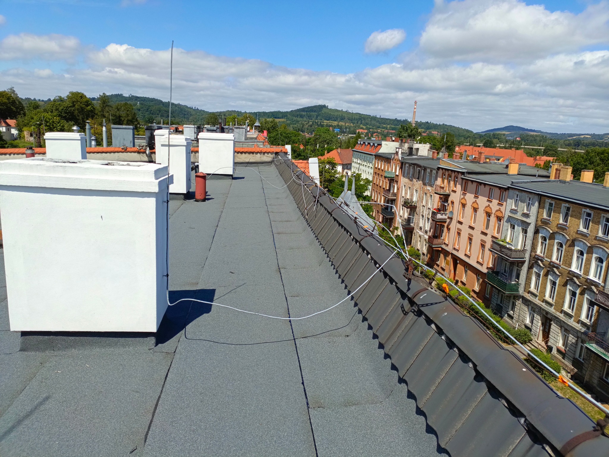 Widok z góry na płaski, szary dach pokryty papą, z białymi kominami i anteną, w tle zabudowa miejska z budynkami o pomarańczowych dachach i zielenią drzew.