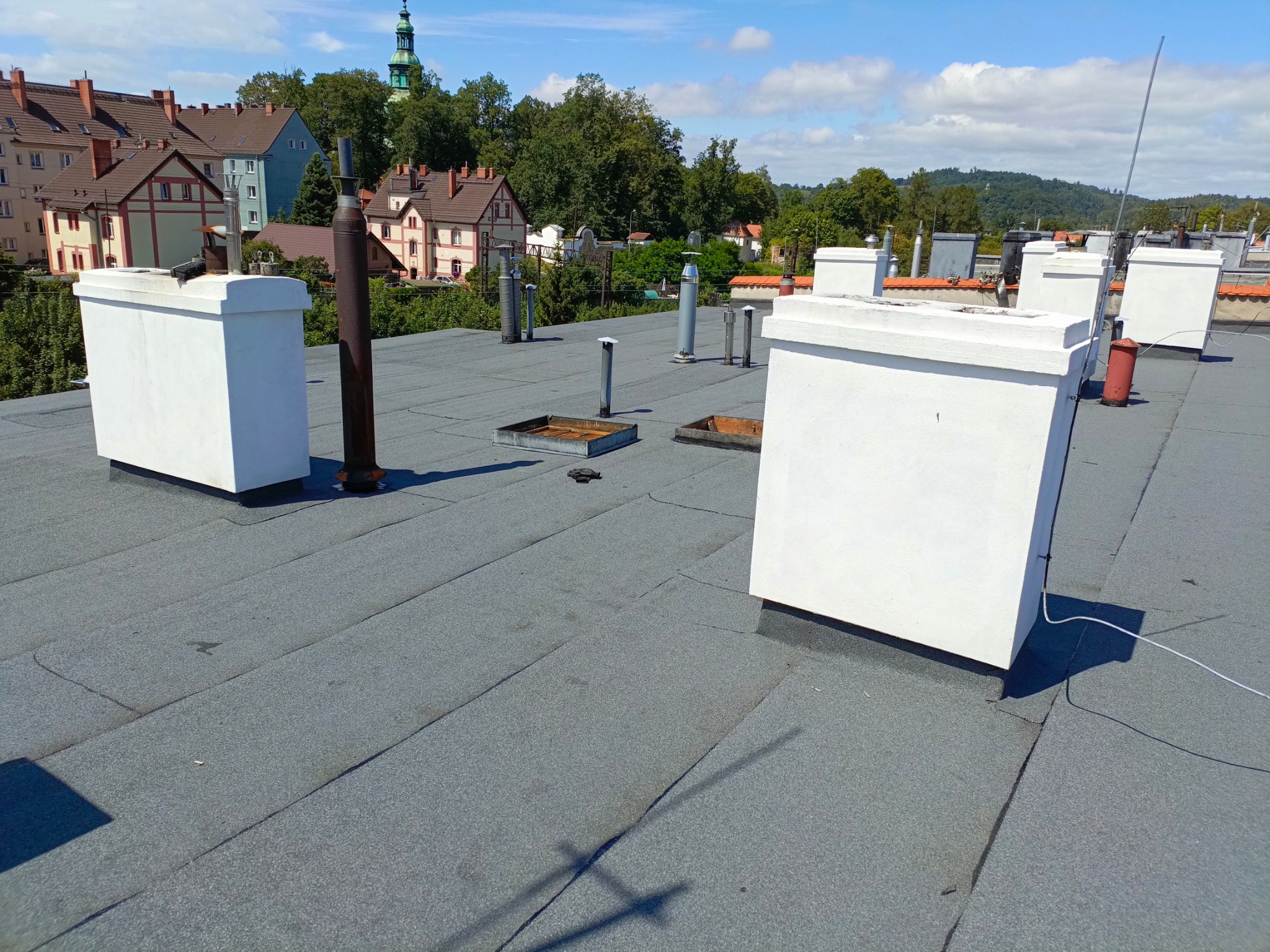 Płaski dach pokryty papą termozgrzewalną z białymi obudowami kominowymi i elementami wentylacyjnymi na tle zabudowy miejskiej z wieżą kościelną w oddali.