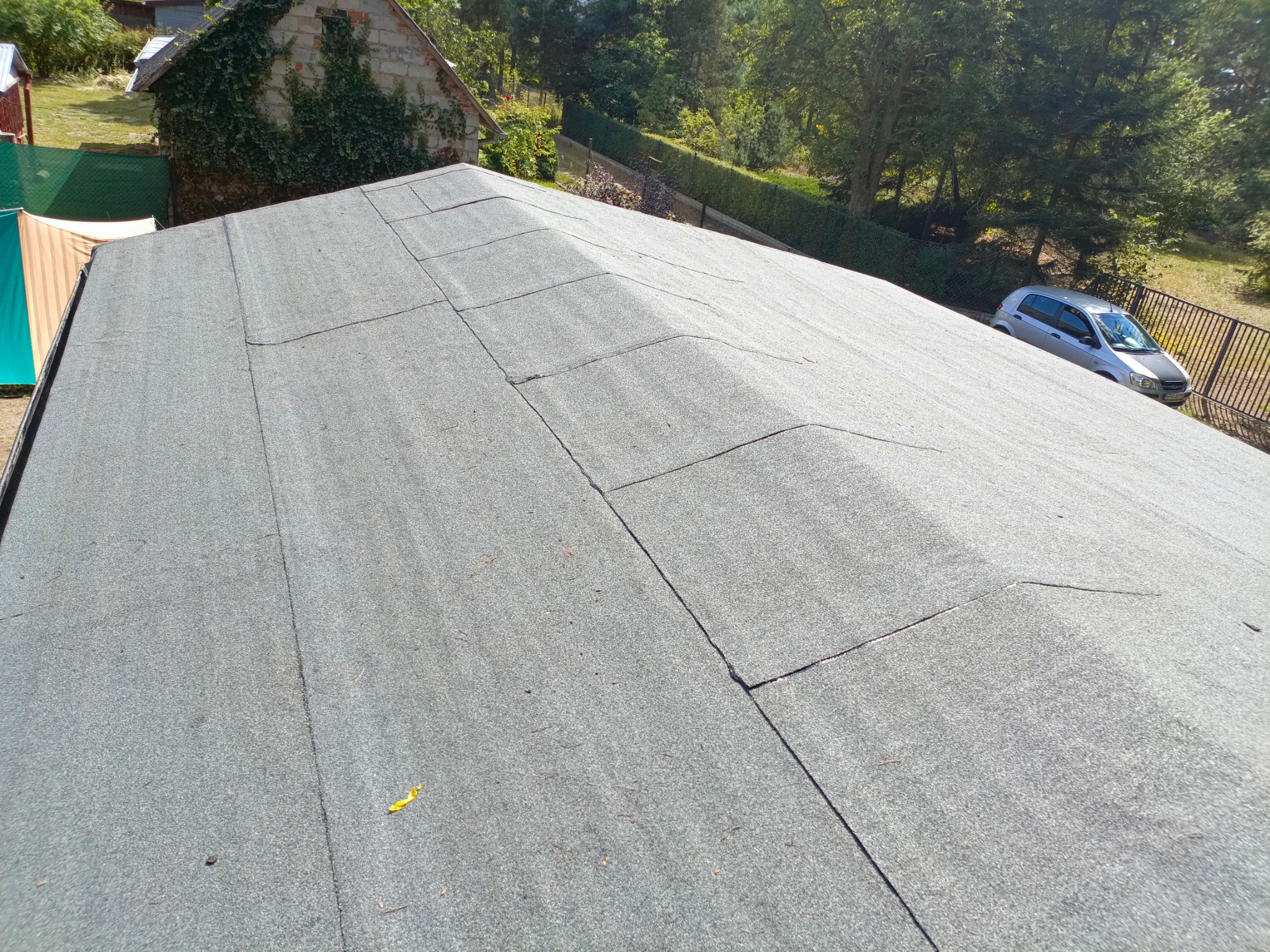 Dach pokryty papą termozgrzewalną, widok z góry, w tle dom z porośniętą bluszczem ścianą i srebrny samochód osobowy.