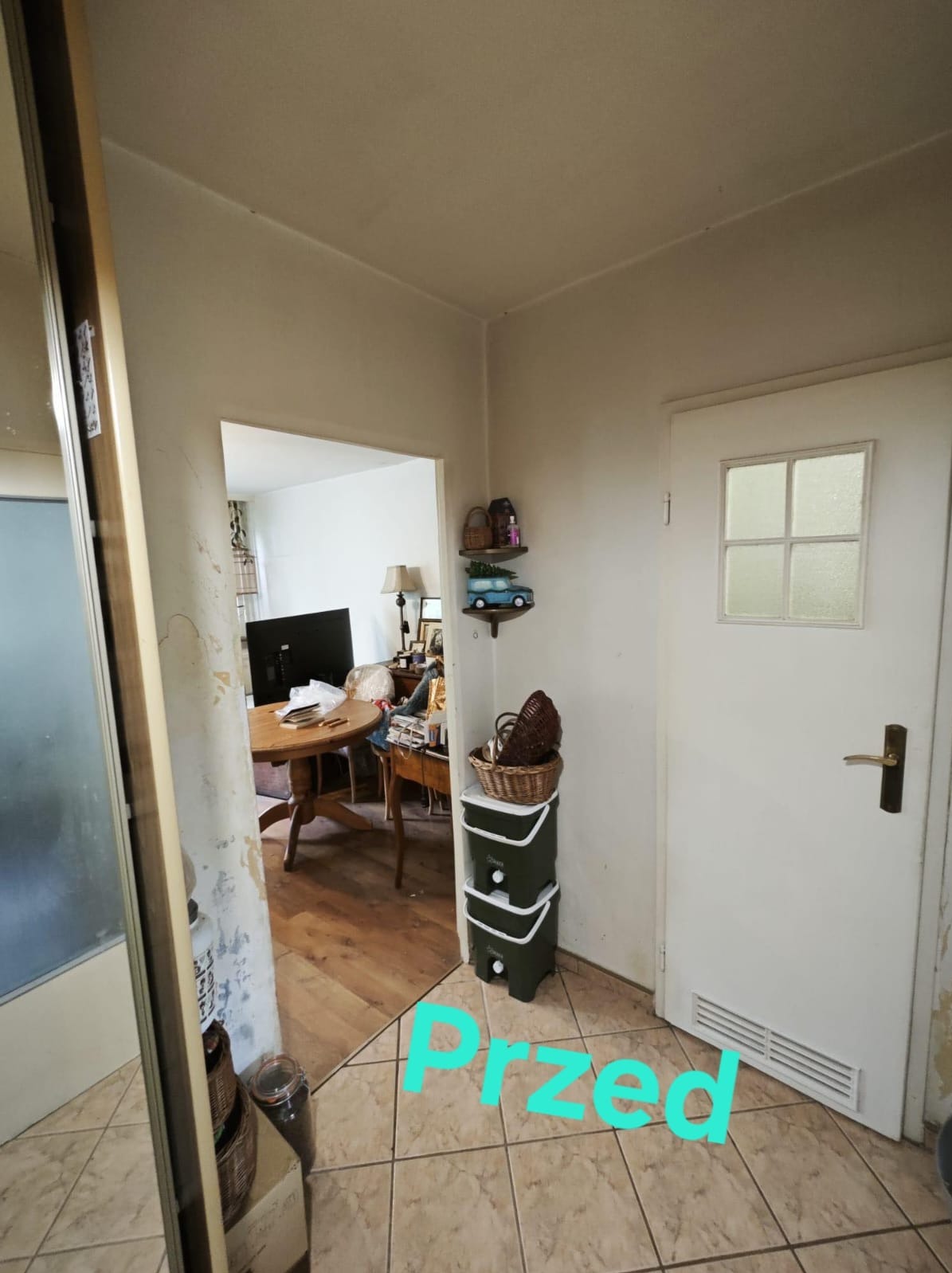 Korytarz mieszkania przed remontem: ściany z widocznymi śladami po usuniętej tapecie, stara podłoga z płytek, widoczna część salonu z drewnianym stołem i telewizorem, białe drzwi z małym okienkiem.