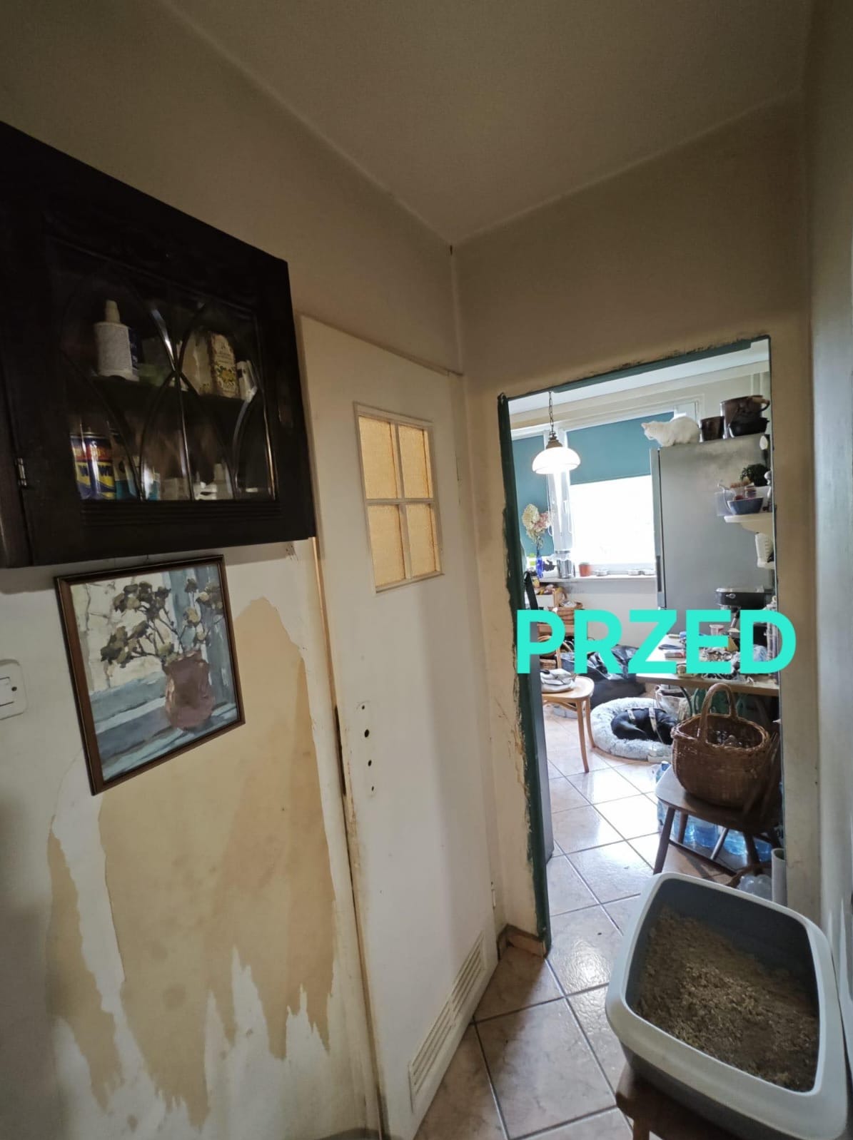 Korytarz we Wrocławiu przed remontem: ściany z widocznymi śladami wilgoci, stara szafka z przeszklonymi drzwiczkami, obrazek na ścianie i widok na zagracone pomieszczenie przez otwarte drzwi.