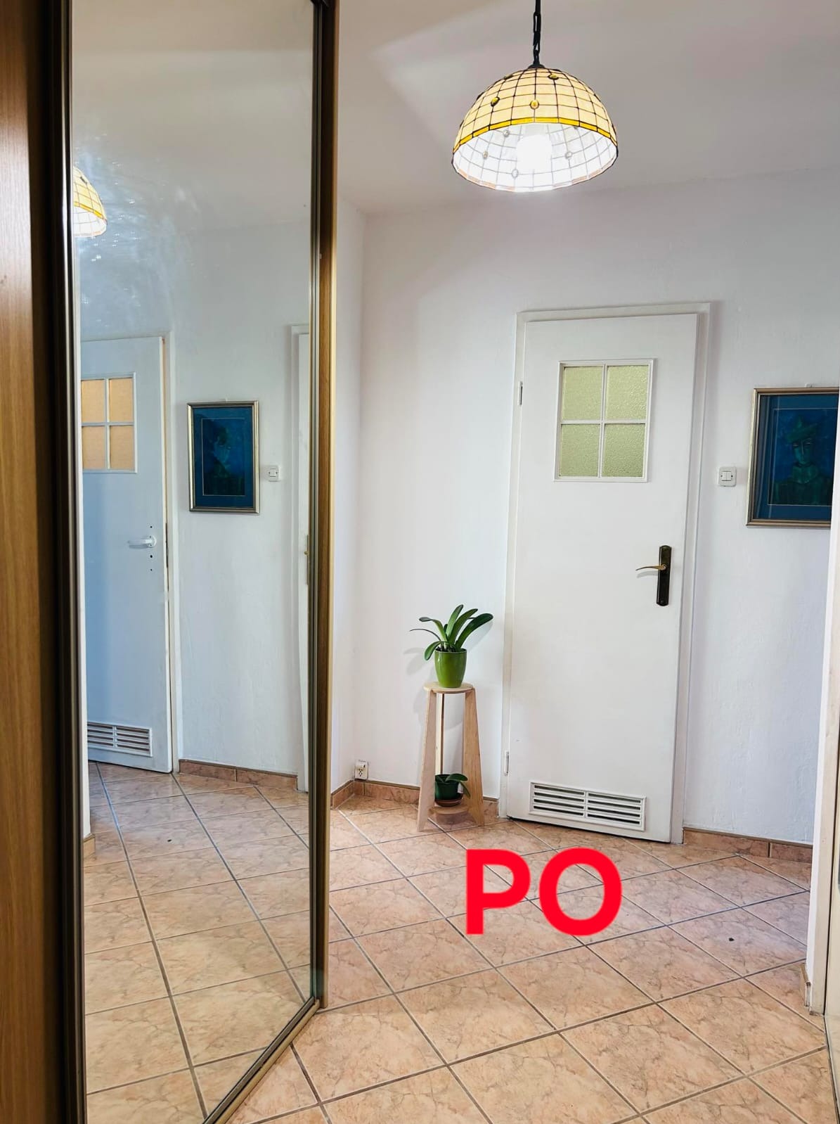 Przedpokój z lustrzaną szafą przesuwną, beżową terakotą, białymi ścianami i drzwiami z szybką, oświetlony lampą w stylu Tiffany, z roślinami na drewnianym stoliku.