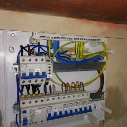 ELEKTOM Tomasz Czopek - Rozdzielnica elektryczna Schrack zamontowana na ścianie, widoczne bezpieczniki, przewody w kolorach niebieskim, żółto-zielonym i brązowym, instalacja w trakcie podłączania.