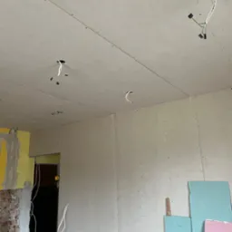 Wnętrze w trakcie remontu: zamontowane płyty karton-gips na ścianach i suficie, widoczne kable elektryczne wystające z otworów w suficie, fragment odsłoniętej cegły i resztki żółtej farby...