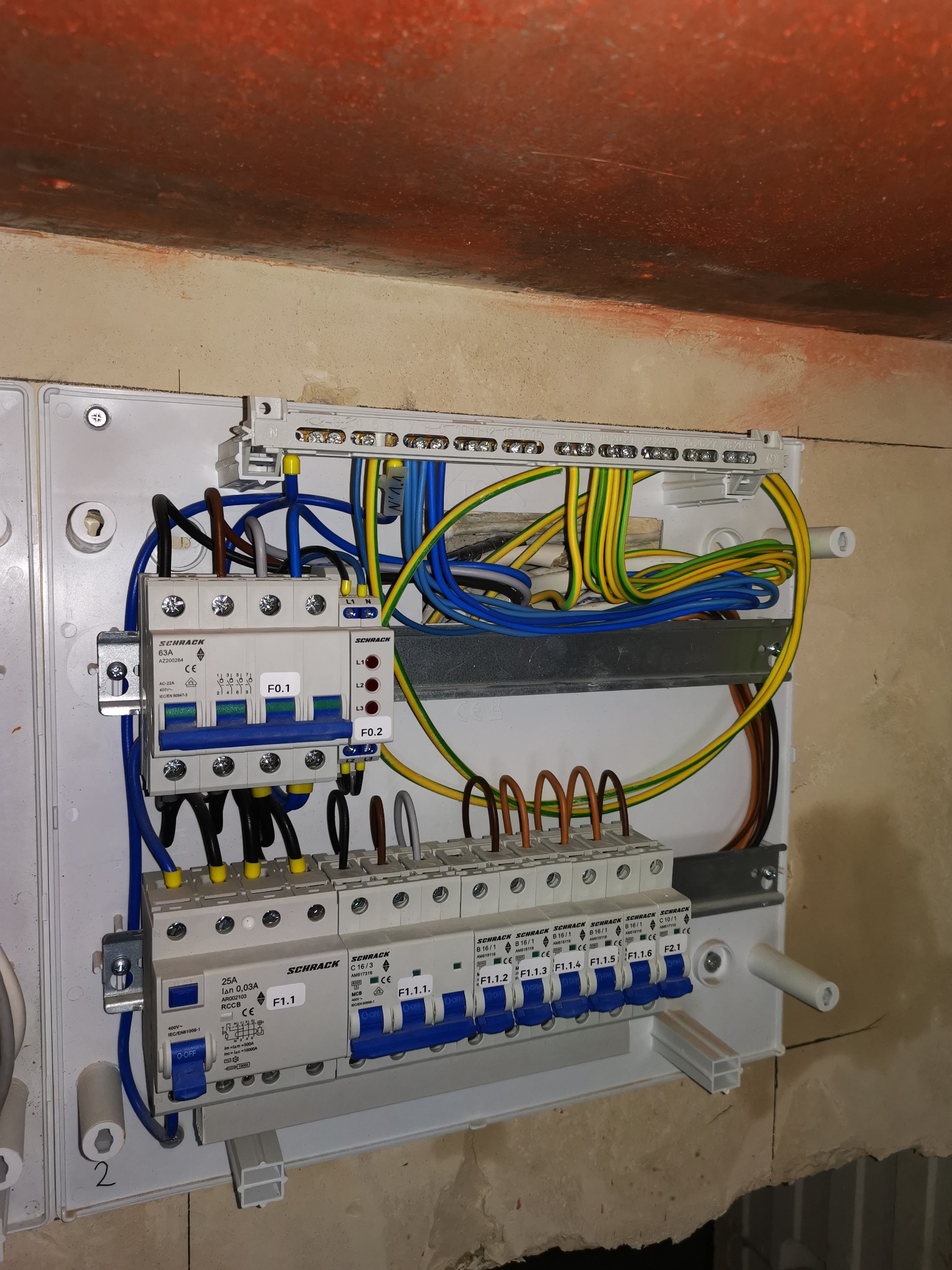 Rozdzielnica elektryczna Schrack zamontowana na ścianie, widoczne bezpieczniki, przewody w kolorach niebieskim, żółto-zielonym i brązowym, instalacja w trakcie podłączania.