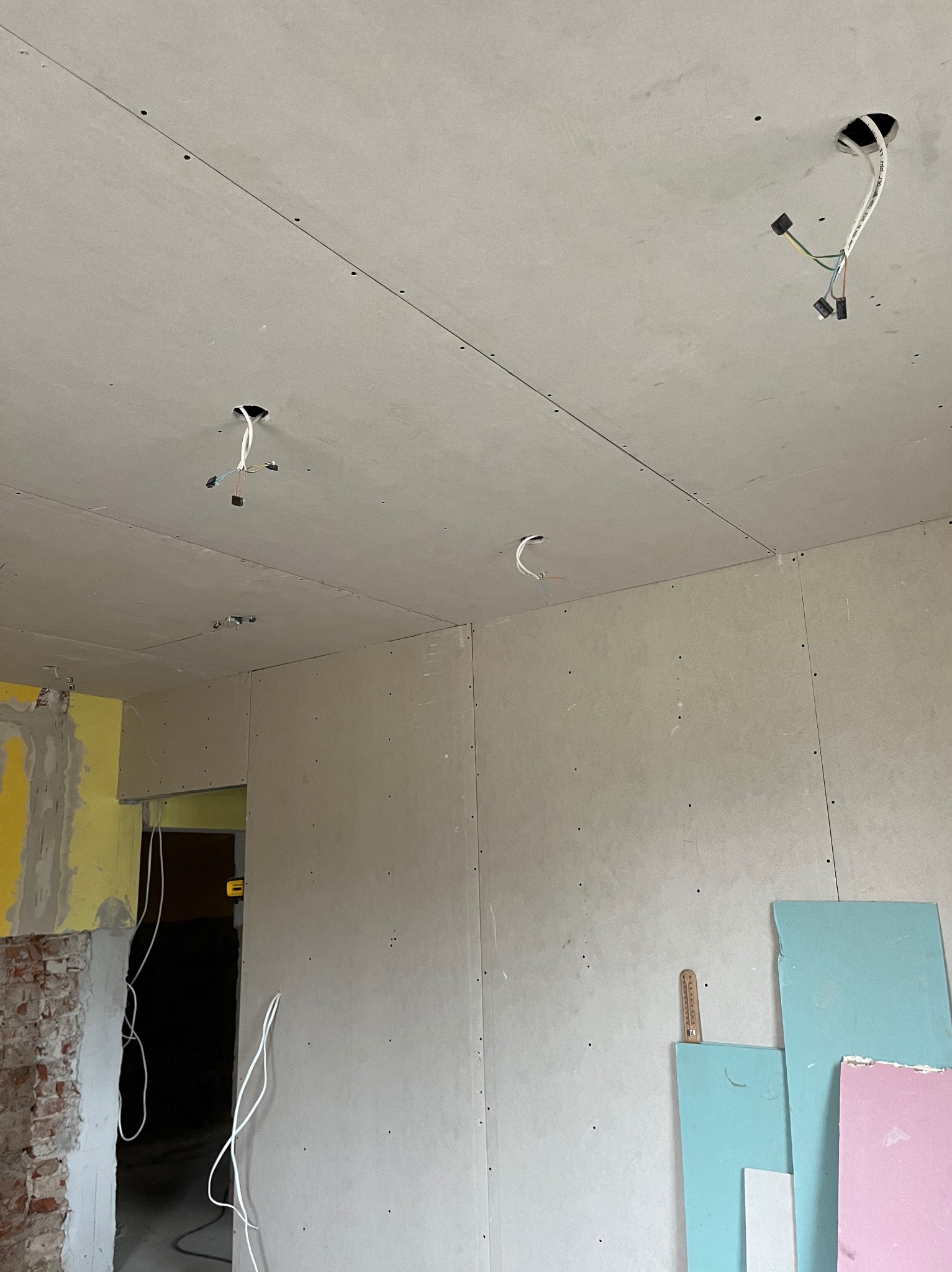 Wnętrze w trakcie remontu: zamontowane płyty karton-gips na ścianach i suficie, widoczne kable elektryczne wystające z otworów w suficie, fragment odsłoniętej cegły i resztki żółtej farby...
