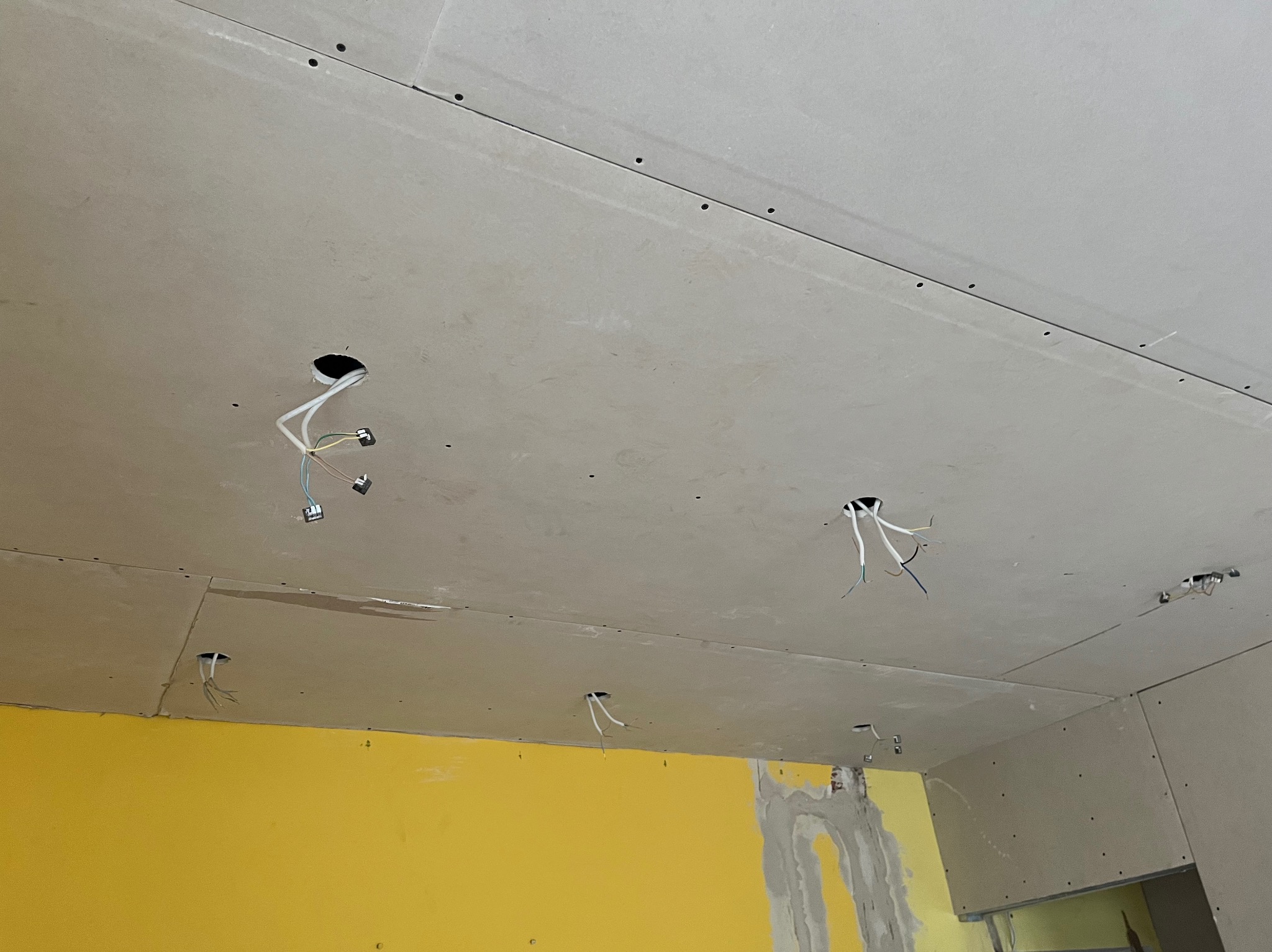 Sufit z płyt kartonowo-gipsowych z widocznymi otworami na instalację elektryczną, wystające przewody, żółta ściana w tle, widoczne łączenia płyt.