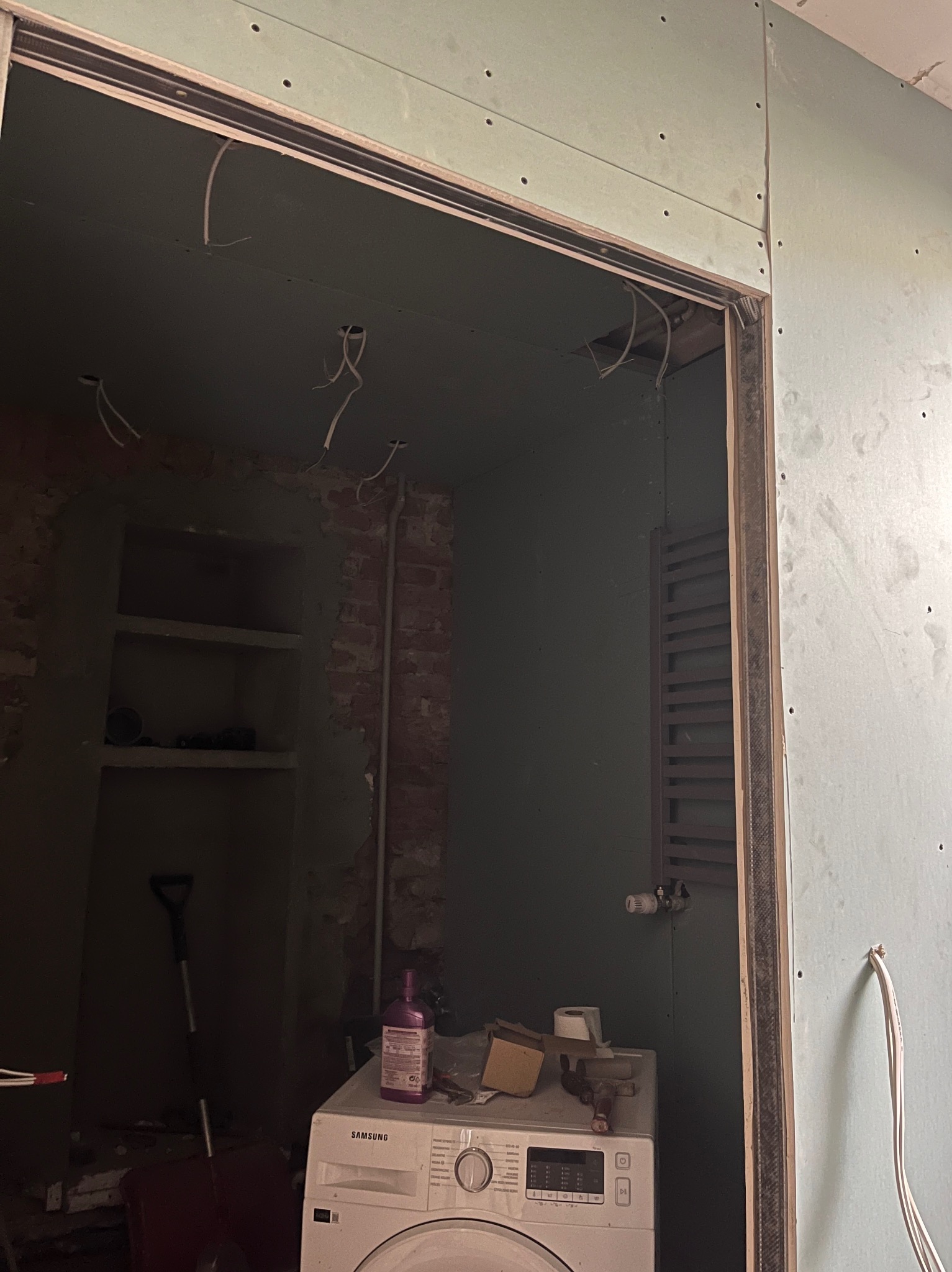Łazienka w trakcie remontu: widoczne płyty kartonowo-gipsowe na ścianach, wystające przewody elektryczne z sufitu, pralka oraz grzejnik drabinkowy w tle, ceglana ściana z wnęką.