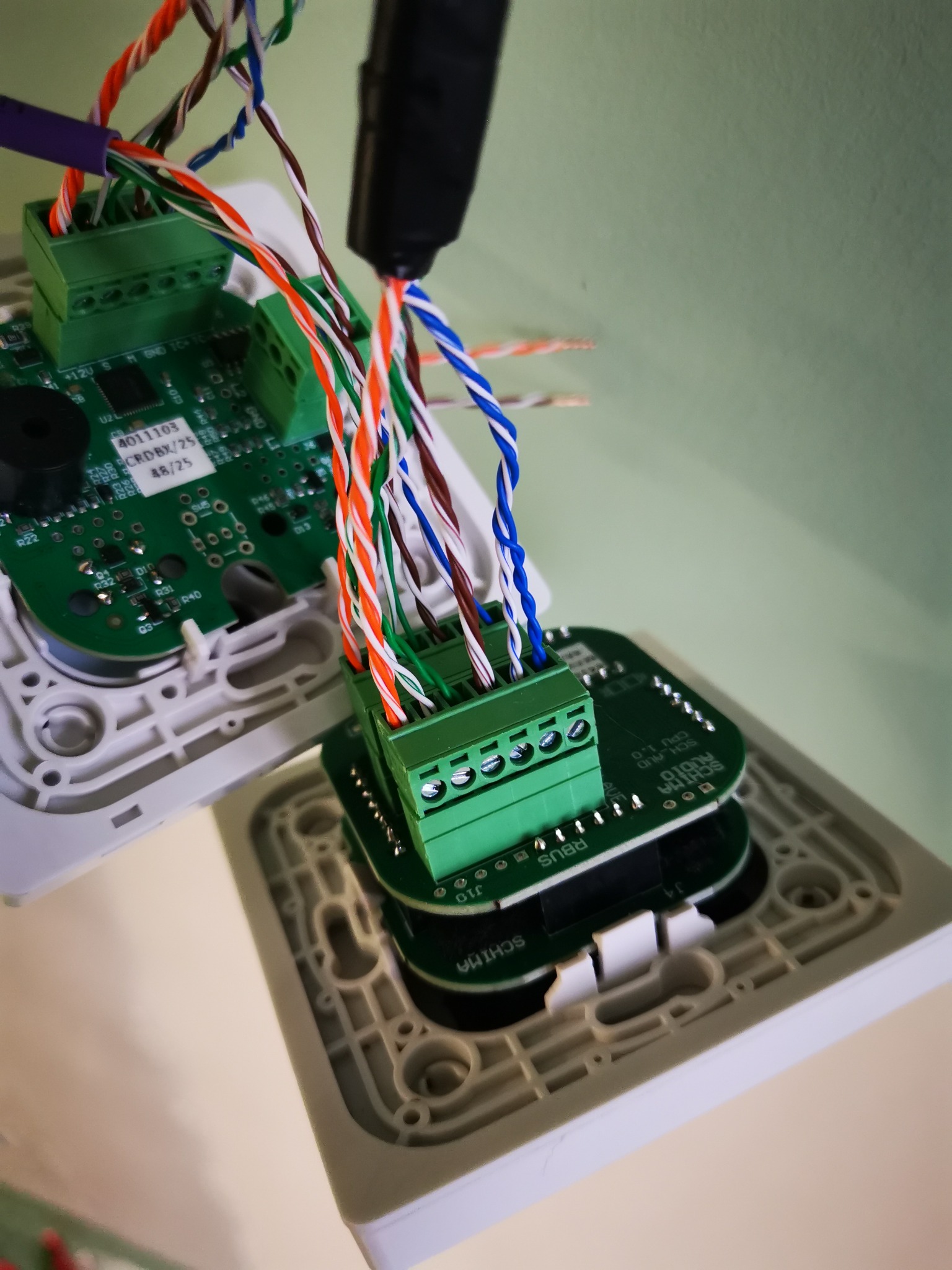 Podłączenie przewodów do płyty sterującej domofonu. Widoczne kolorowe kable wpięte w zielone złącza śrubowe. Biała obudowa i fragment zielonej ściany w tle.