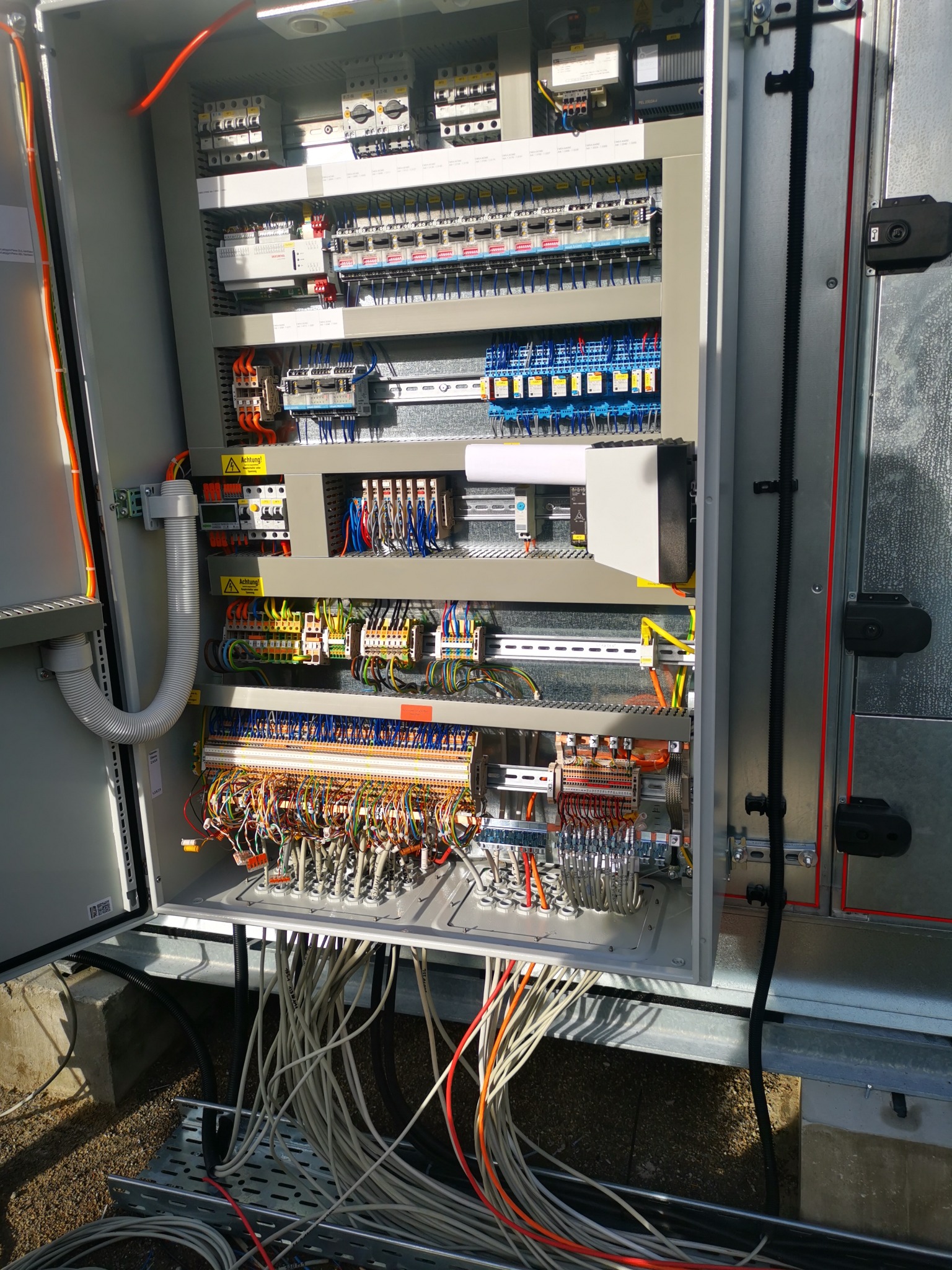 Otwarta szafa sterownicza z licznymi przewodami i elementami elektrycznymi, widoczne połączenia i zabezpieczenia. Szara obudowa, kable w różnych kolorach.