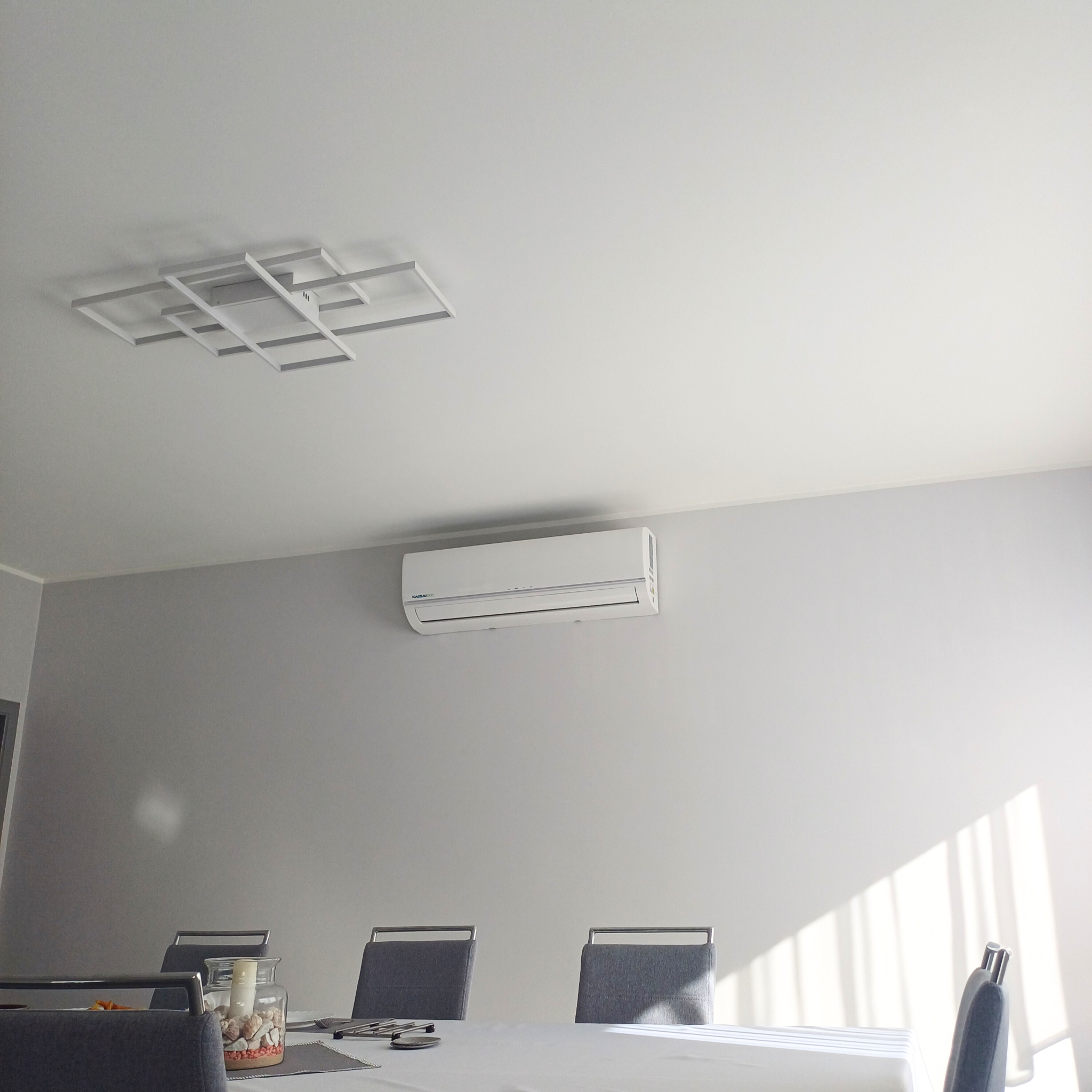 Biały klimatyzator Kaisai Eco zamontowany na szarej ścianie nad stołem w jasnym pomieszczeniu z modernistyczną lampą sufitową.
