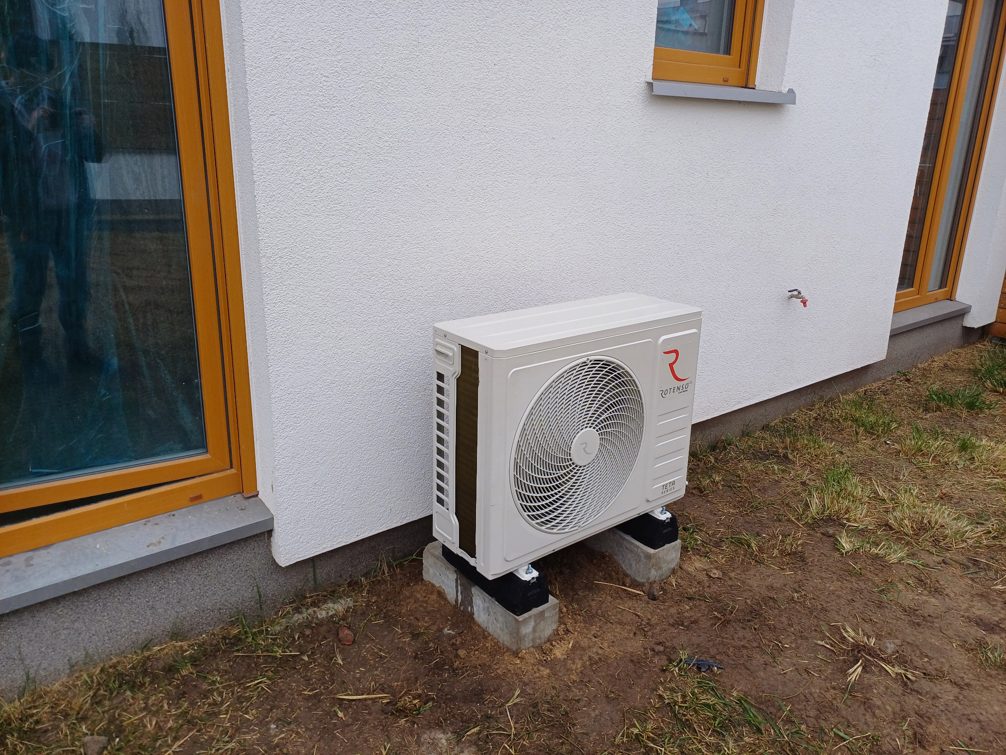 Biała zewnętrzna jednostka klimatyzacji Rotenso Teta Series zamontowana na betonowych wspornikach przy ścianie domu z oknami w drewnianych ramach.