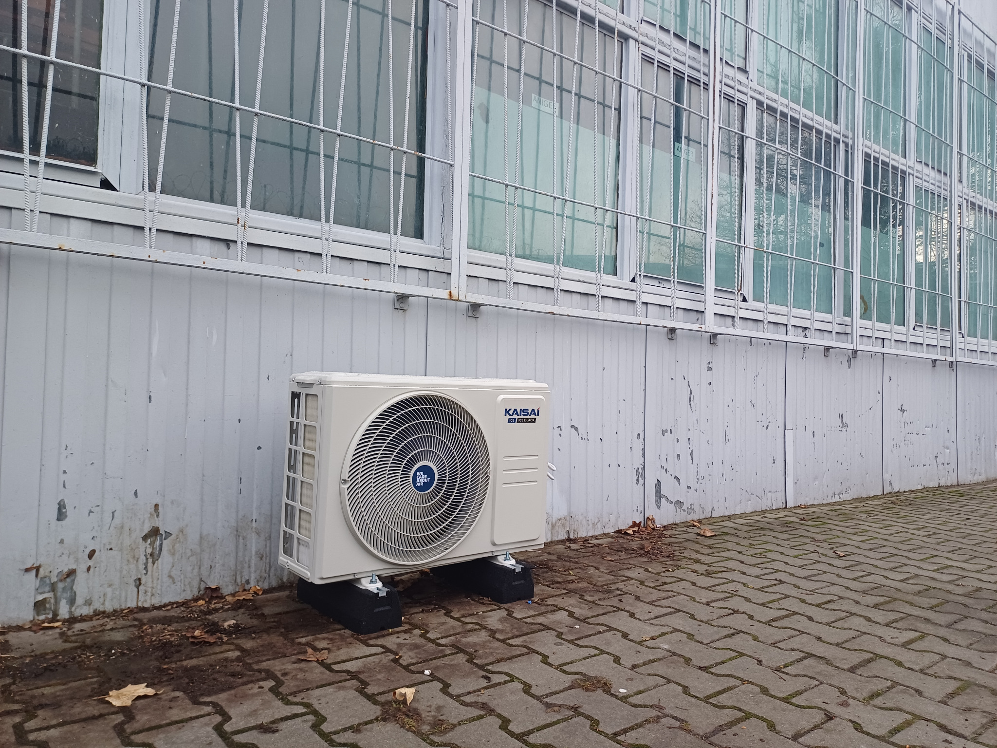 Zewnętrzna jednostka klimatyzacji Kaisai Ice/Ice Black na czarnych podkładkach, zamontowana przy pomalowanej na szaro ścianie budynku z oknami zabezpieczonymi kratami, na chodniku z kostki brukowej.