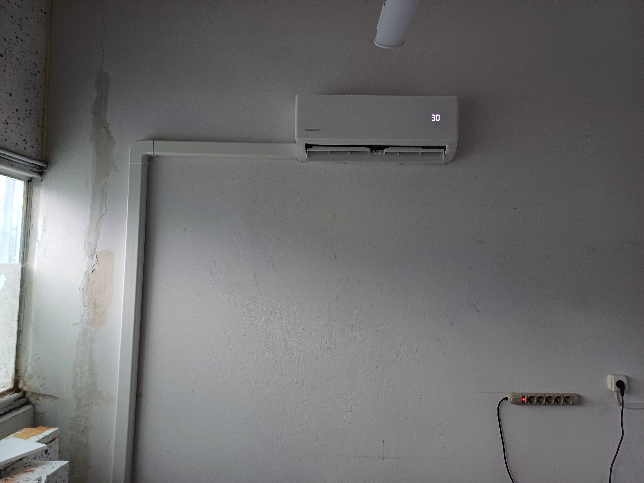 Biała klimatyzacja Kaisai zamontowana na szarej ścianie z widocznymi śladami po naprawach, z podłączoną listwą zasilającą i widoczną lampą sufitową.