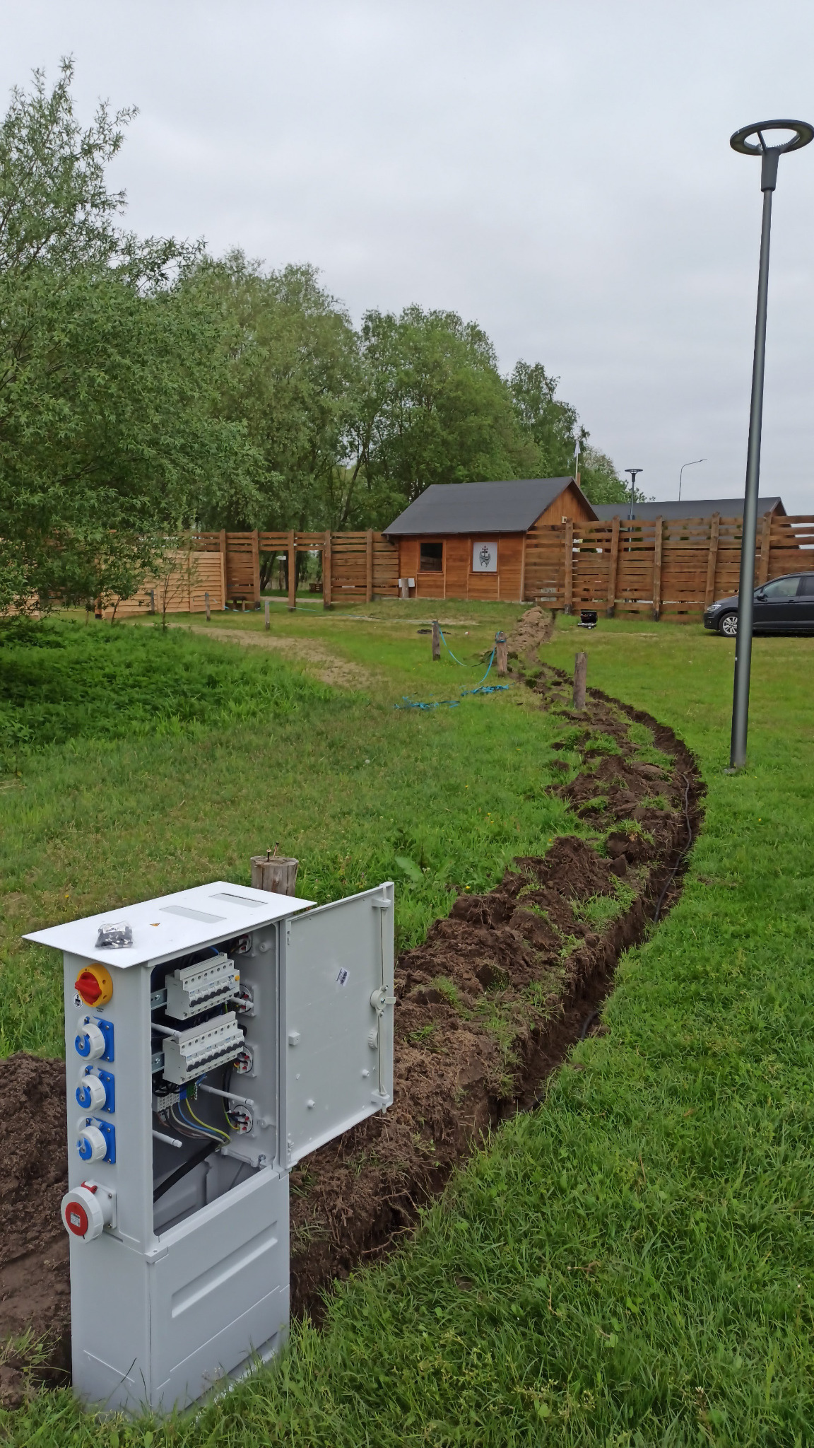 Otwarta szafa elektryczna z gniazdami siłowymi i zabezpieczeniami, umieszczona w wykopie na trawniku, widoczna linia wykopu ciągnąca się w kierunku drewnianego budynku z ogrodzeniem w tle.