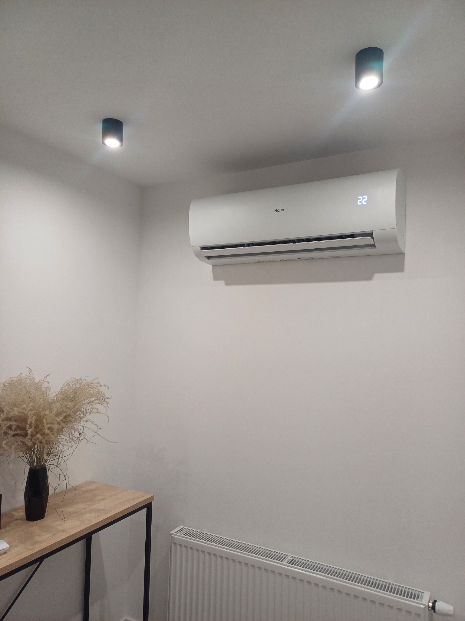 Biała klimatyzacja Haier na ścianie w jasnym pomieszczeniu, ustawiona na 22 stopnie Celsjusza, z widocznym grzejnikiem pod spodem i stolikiem z wazonem z suszonymi kwiatami z boku.