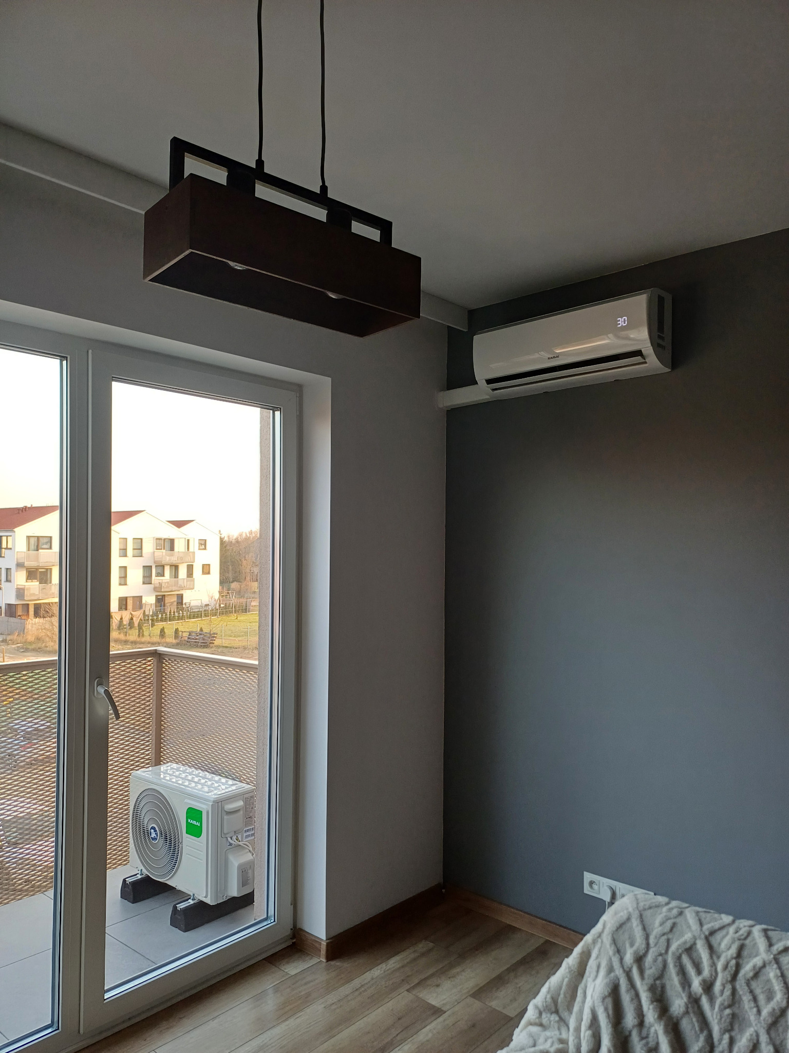 Wnętrze pokoju z zamontowaną klimatyzacją typu split: jednostka wewnętrzna na ścianie, jednostka zewnętrzna na balkonie za przeszklonymi drzwiami balkonowymi, nowoczesna lampa sufitowa.