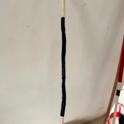 Biały przewód elektryczny z czarną taśmą izolacyjną na tle jasnej ściany z widocznymi zabrudzeniami i elementami wyposażenia wnętrza.