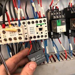 INSTALATORSTWO ELEKTRYCZNE Artur Marański - Elektryk wymienia kondensator 2µF-U 450WV AC wewnątrz szarej metalowej szafy sterowniczej z oznaczonymi kablami i przekaźnikami, widoczne narzędzie.