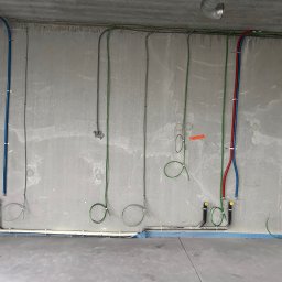 INSTALATORSTWO ELEKTRYCZNE Artur Marański - Surowa ściana z widocznymi instalacjami elektrycznymi i wodno-kanalizacyjnymi w trakcie montażu w Jasionowie. Widoczne kable i rury prowadzone po ścianie, niektóre zakończone zaślepkami.