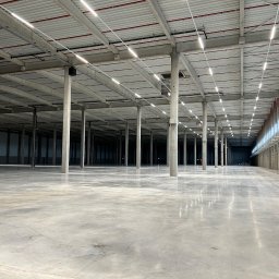 INSTALATORSTWO ELEKTRYCZNE Artur Marański - Wnętrze hali magazynowej z betonową posadzką i rzędami kolumn wspierających sufit z widocznymi liniami oświetlenia LED.