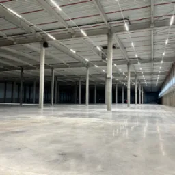 Wnętrze hali magazynowej z betonową posadzką i rzędami kolumn wspierających sufit z widocznymi liniami oświetlenia LED.