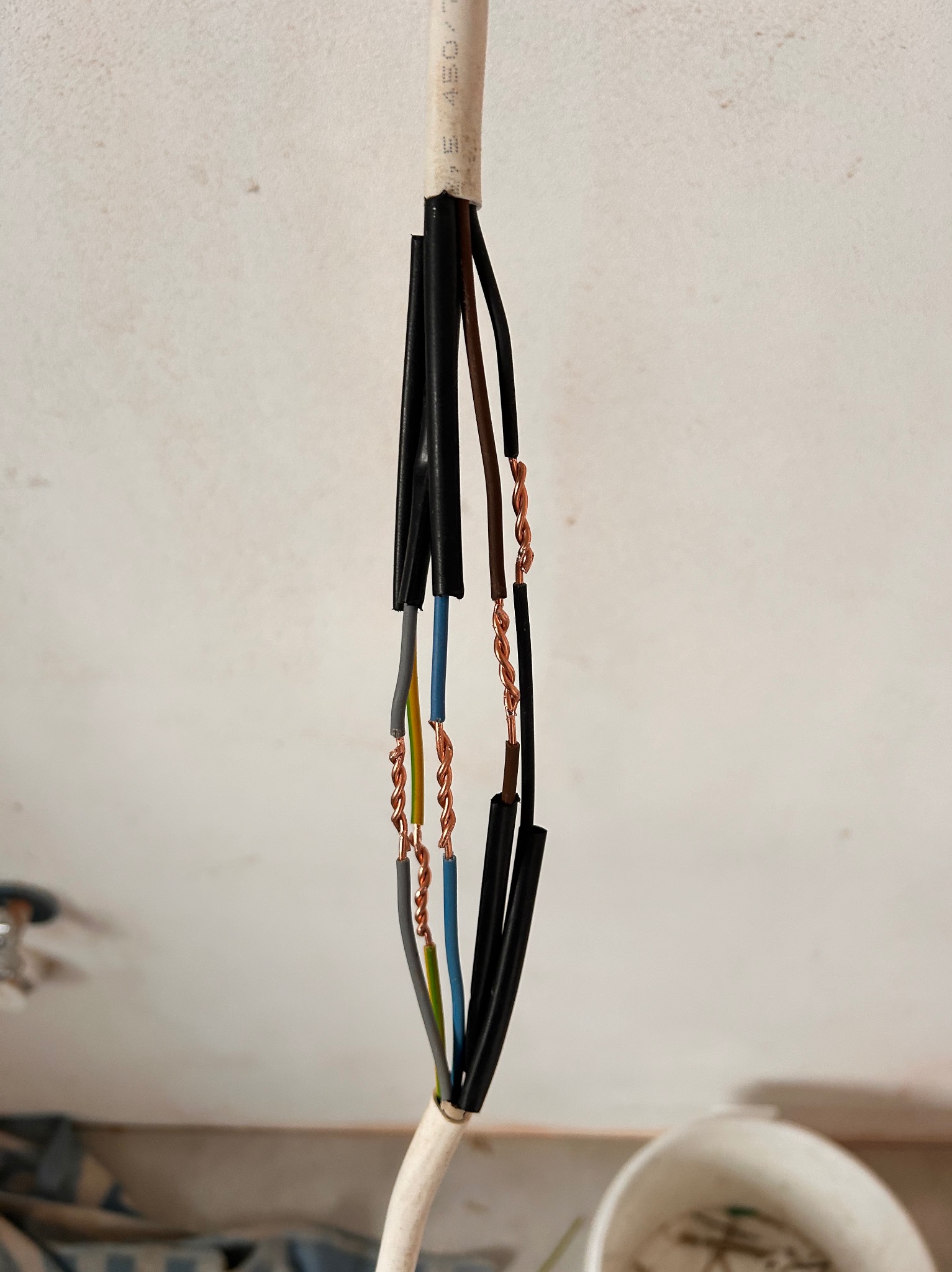 Rozizolowany kabel elektryczny z odsłoniętymi i skręconymi żyłami miedzianymi, wiszący na tle białej ściany.