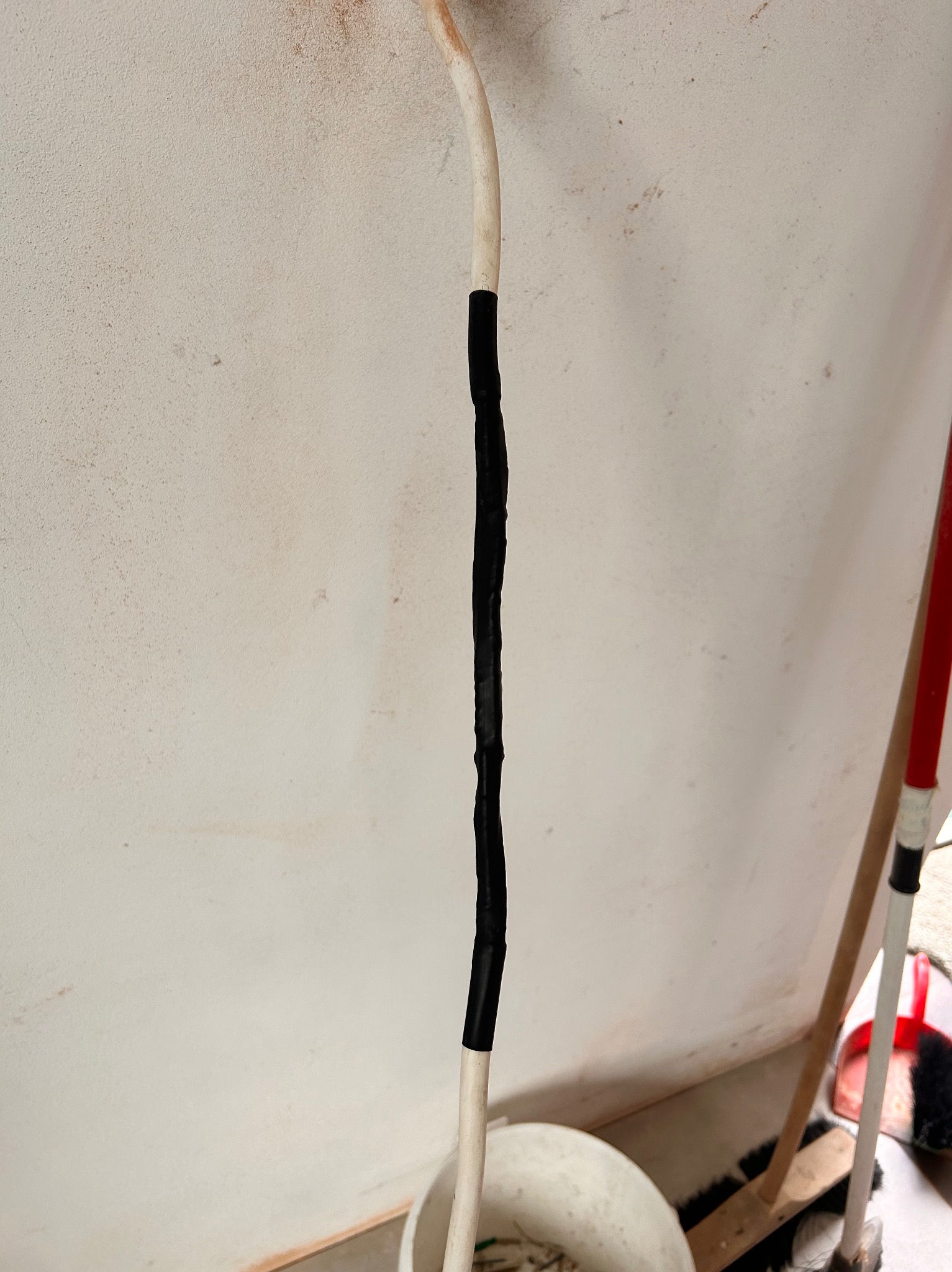 Biały przewód elektryczny z czarną taśmą izolacyjną na tle jasnej ściany z widocznymi zabrudzeniami i elementami wyposażenia wnętrza.