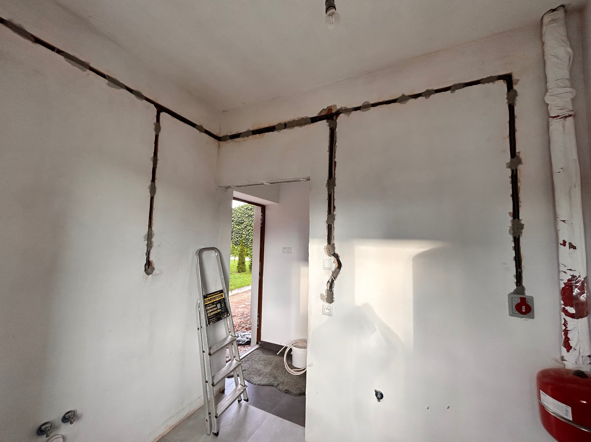 Wnętrze w trakcie remontu z nową instalacją elektryczną prowadzoną po ścianach, zabezpieczoną szarym cementem, widoczna drabina aluminiowa.
