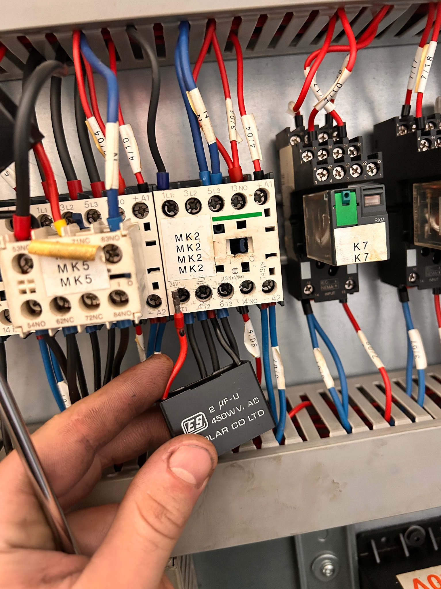 Elektryk wymienia kondensator 2µF-U 450WV AC wewnątrz szarej metalowej szafy sterowniczej z oznaczonymi kablami i przekaźnikami, widoczne narzędzie.