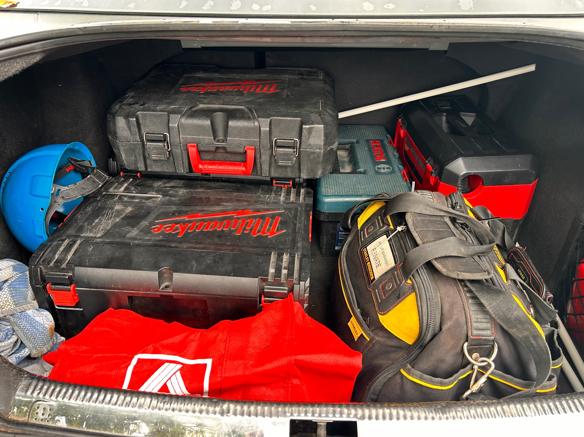 Bagażnik samochodu wypełniony skrzynkami narzędziowymi Milwaukee i Bosch, torbą narzędziową Stanley oraz kaskiem ochronnym, sugerujący gotowość do pracy elektryka.