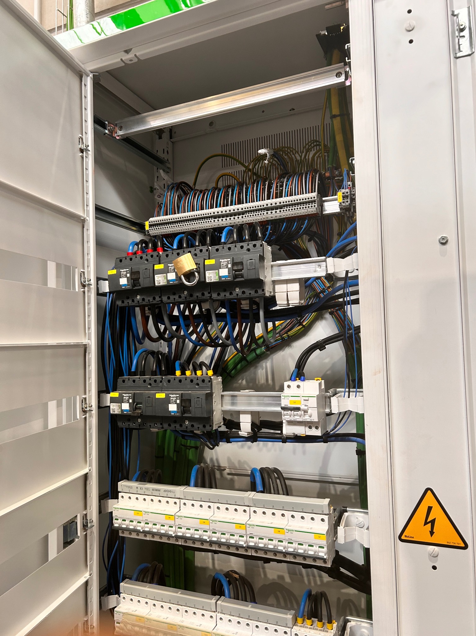 Otwarta szafa elektryczna z widocznymi bezpiecznikami, przewodami i żółtą naklejką ostrzegawczą z symbolem błyskawicy, zabezpieczona kłódką na jednym z wyłączników.