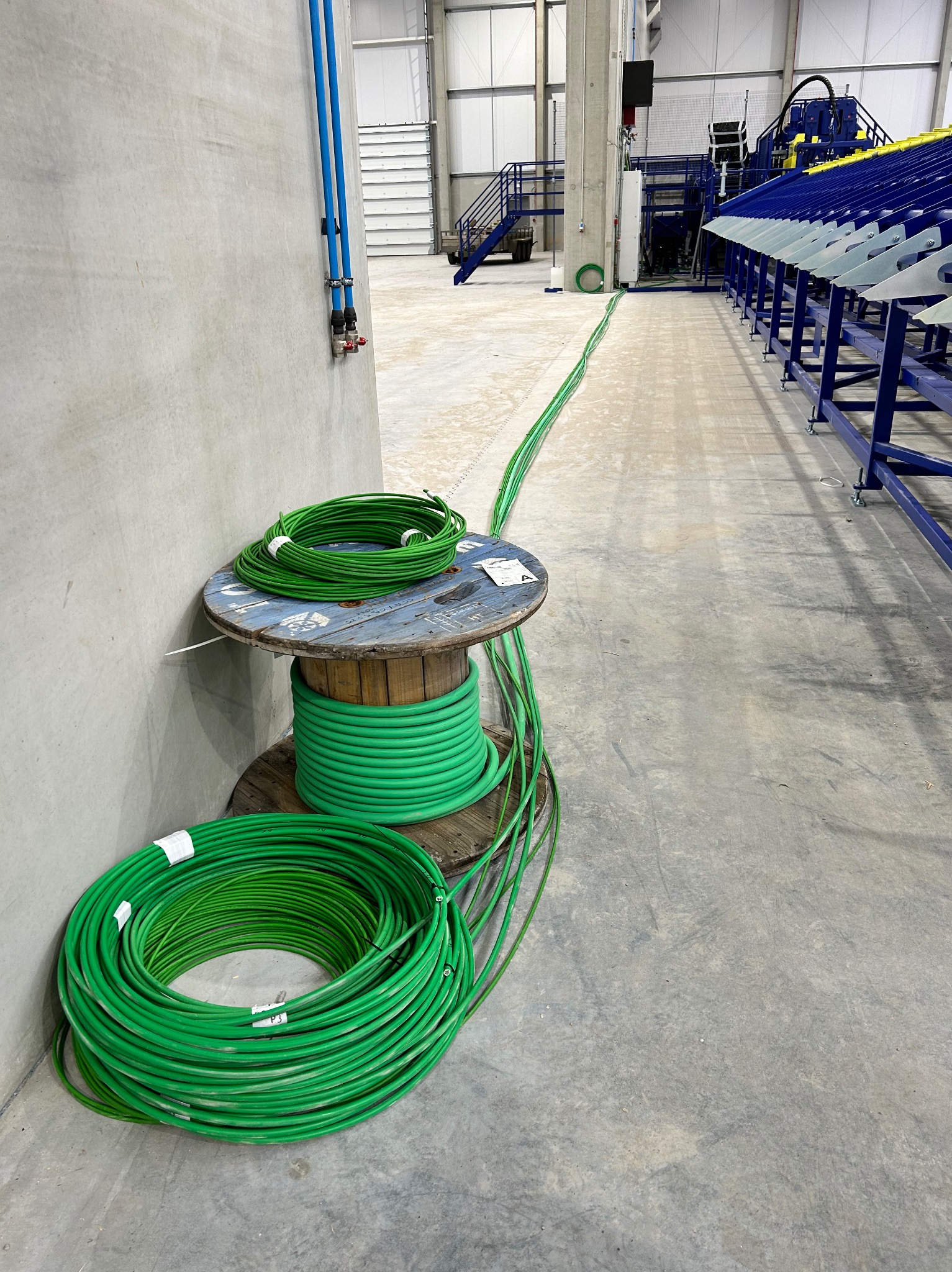 Szpula z nawiniętym zielonym przewodem elektrycznym w hali przemysłowej, przewód rozciągnięty wzdłuż ściany, widoczne niebieskie rury instalacyjne na ścianie.