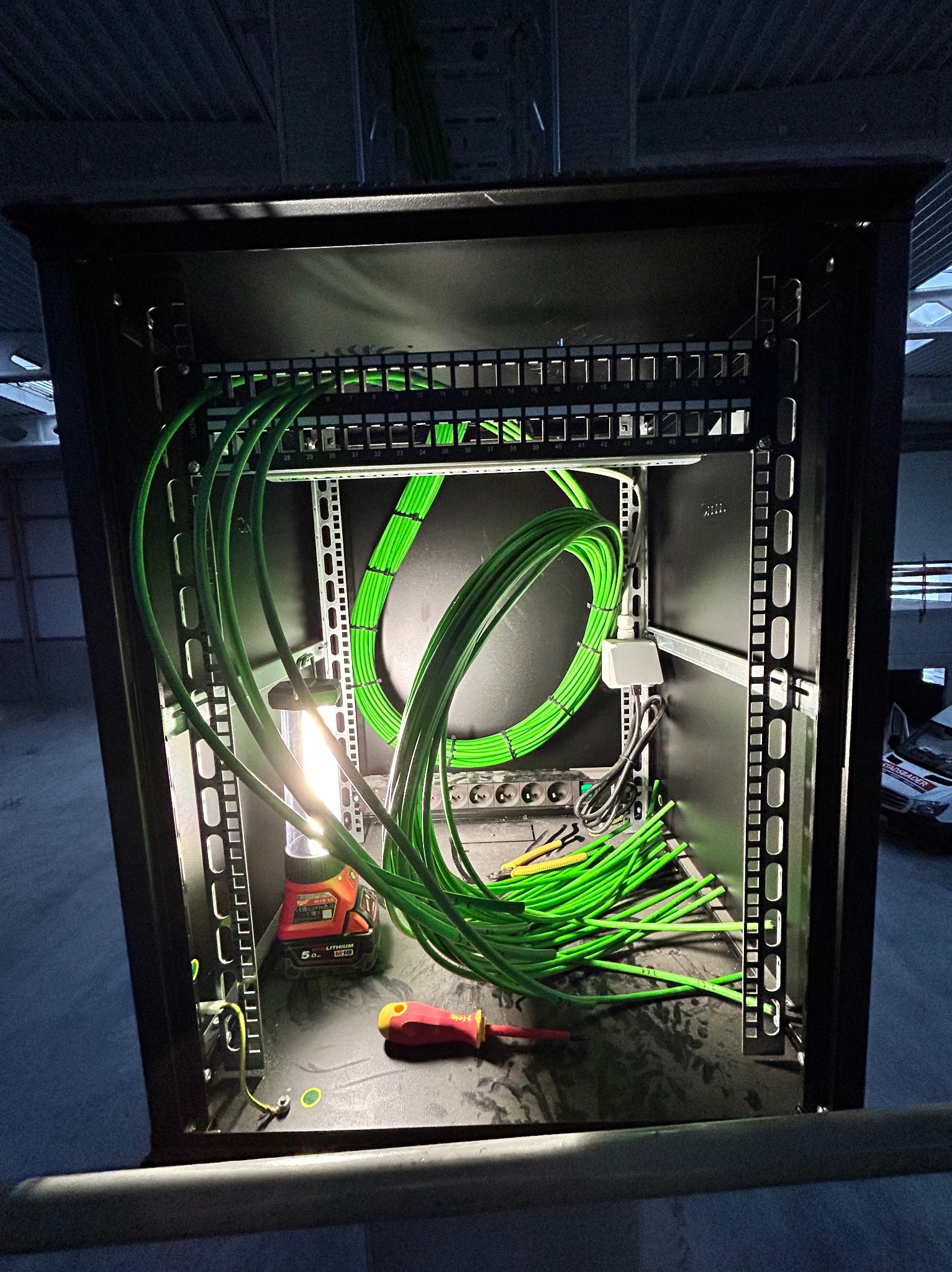 Wnętrze szafy rack z uporządkowanymi zielonymi kablami, oświetlone lampą warsztatową; widoczne narzędzia, w tle fragment pomieszczenia.